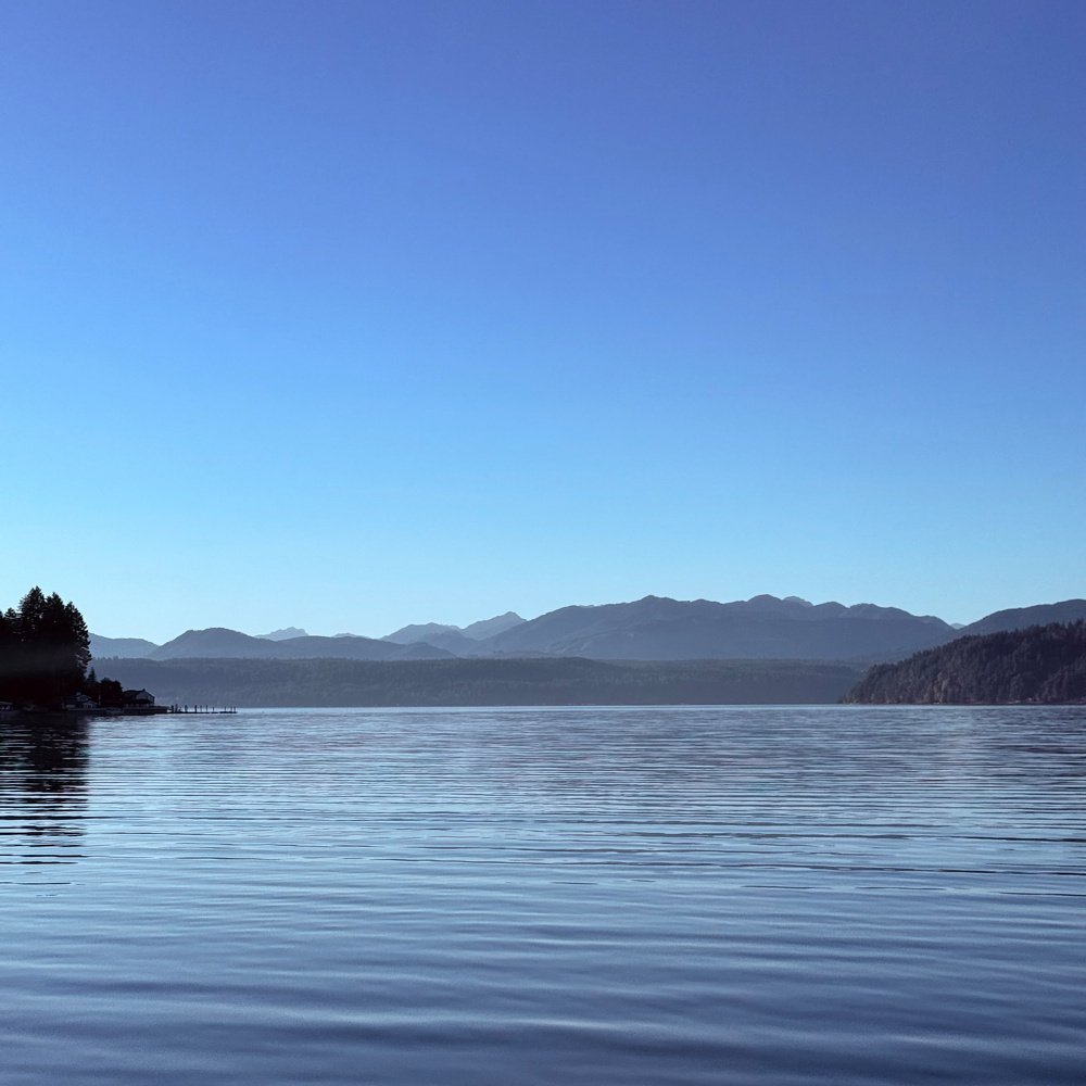 A calm body of water is surrounded by distant mountains under a clear blue sky.