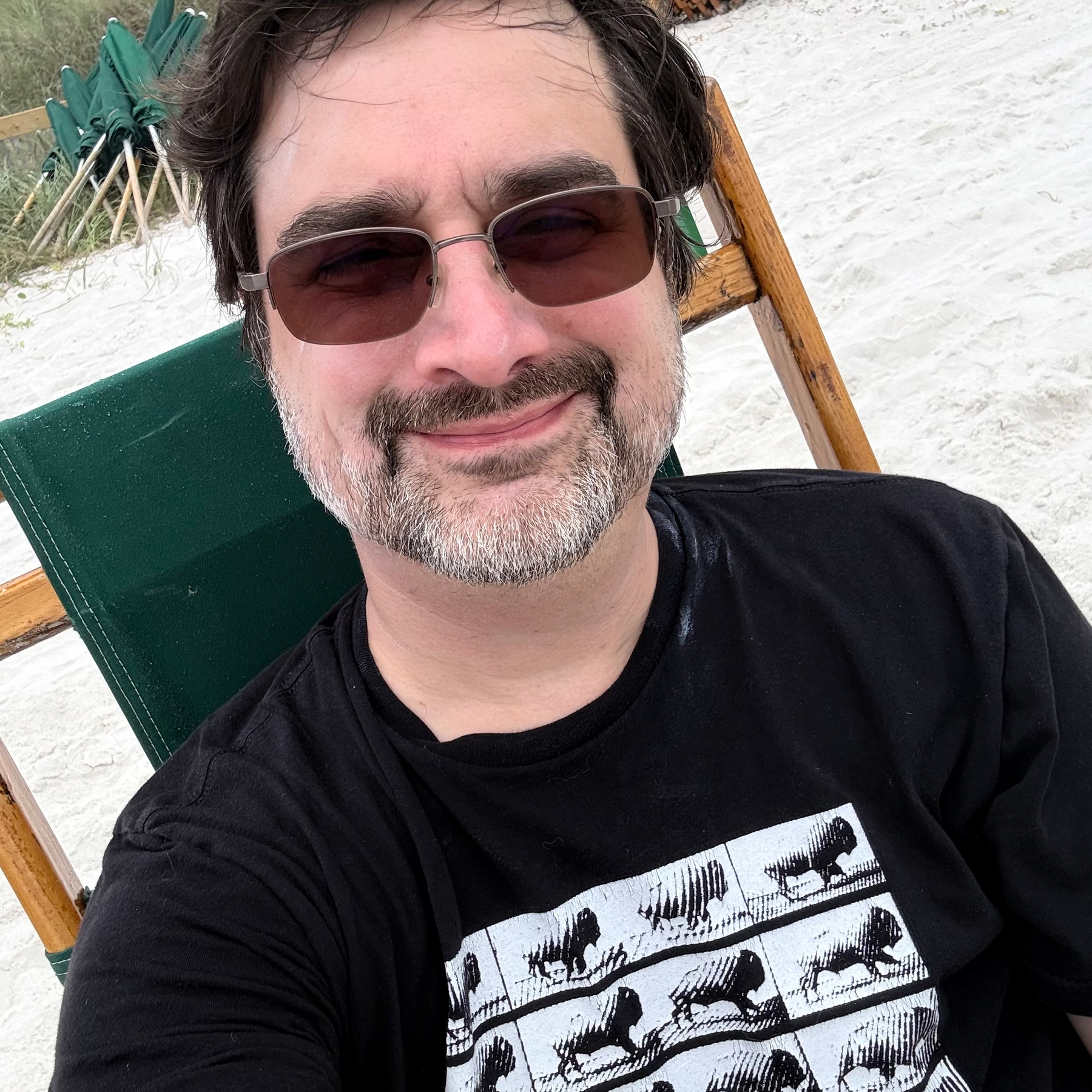 A man with glasses and a beard is smiling while sitting on a green chair on a sandy beach.