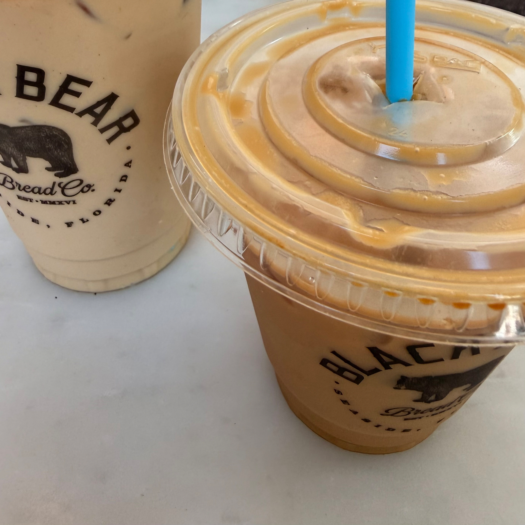 Two iced coffee drinks with Black Bear Bread Co. logos are placed on a light-colored surface, one with a blue straw.