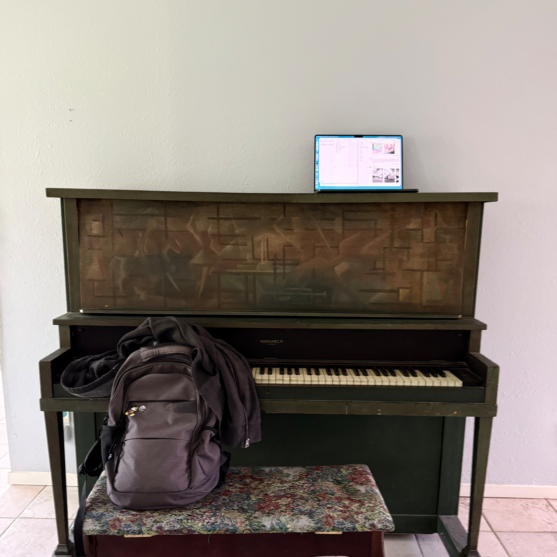 A closed, dark upright piano with a backpack on a bench in front and a laptop resting on top.