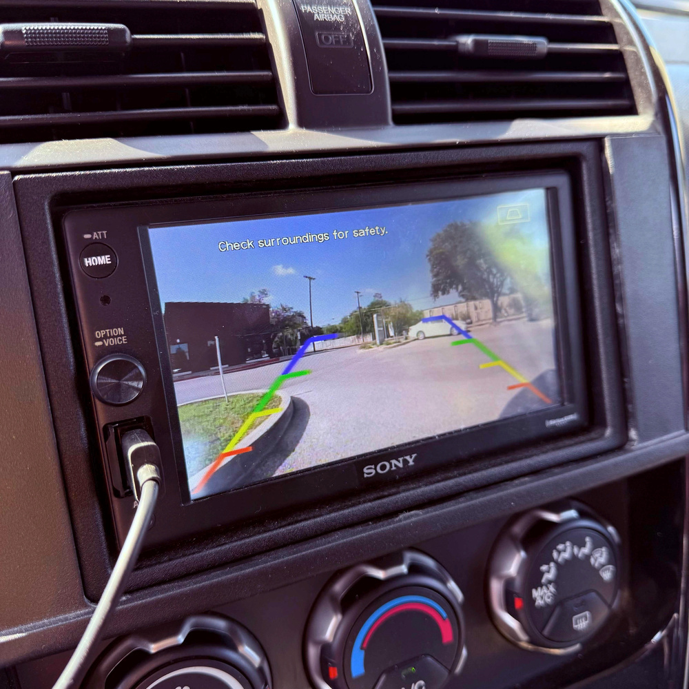 A car dashboard features a Sony screen displaying the rearview camera with parking guidelines and a warning to check surroundings for safety.