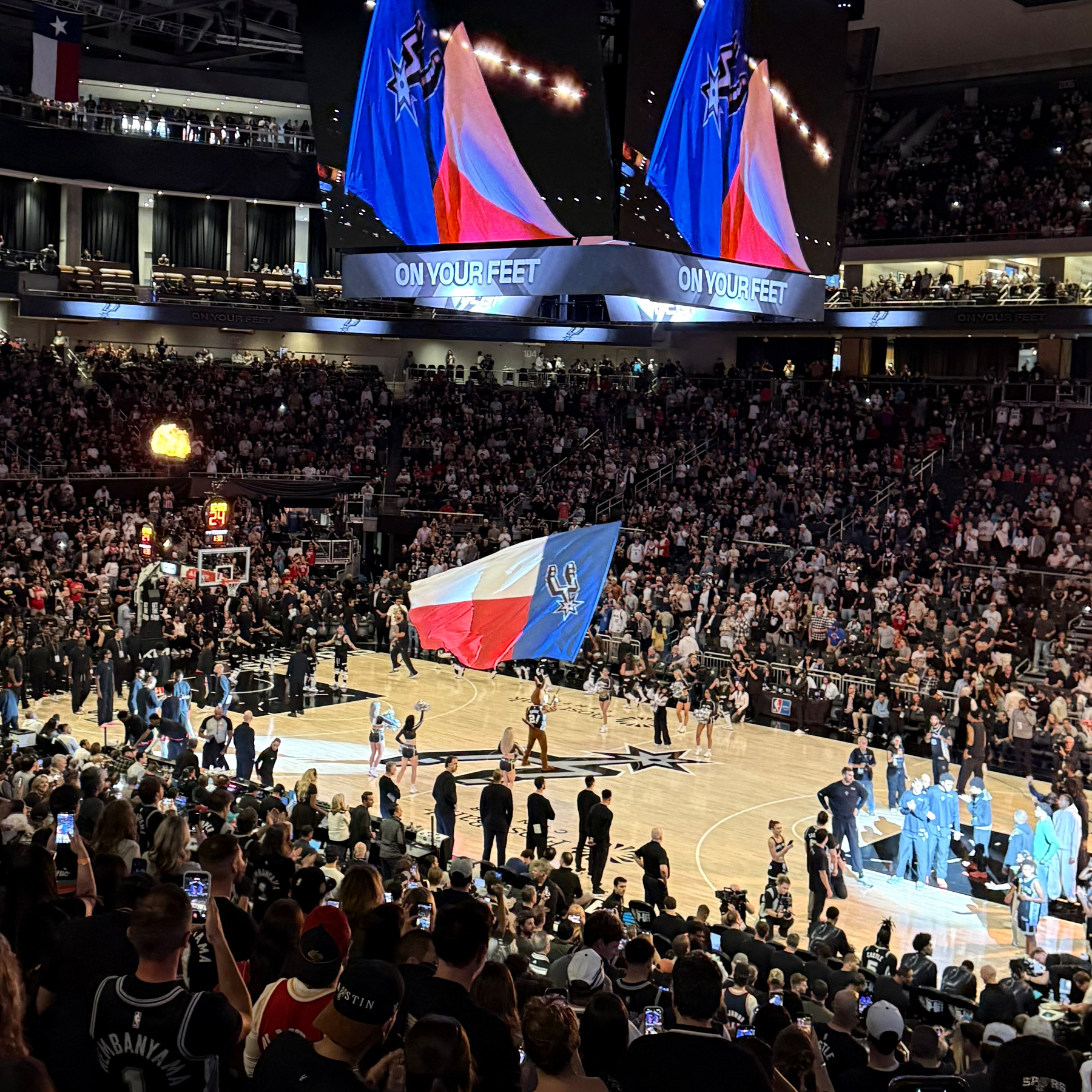 A large crowd is gathered in Moody Center for a basketball game with the Coyote on the court waving a Texas state flag with the Spurs logo.