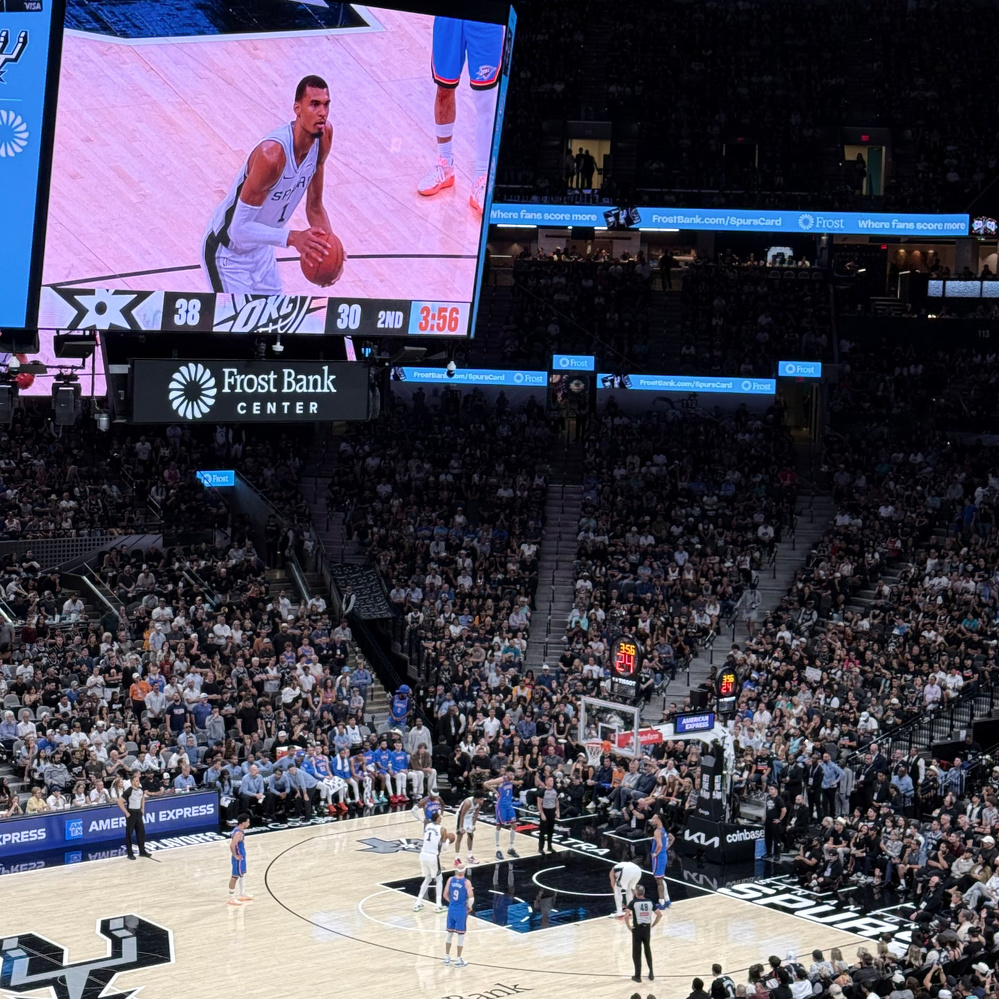 A basketball game is being played in an indoor arena with a large screen displaying the score and Wemby taking a free throw.