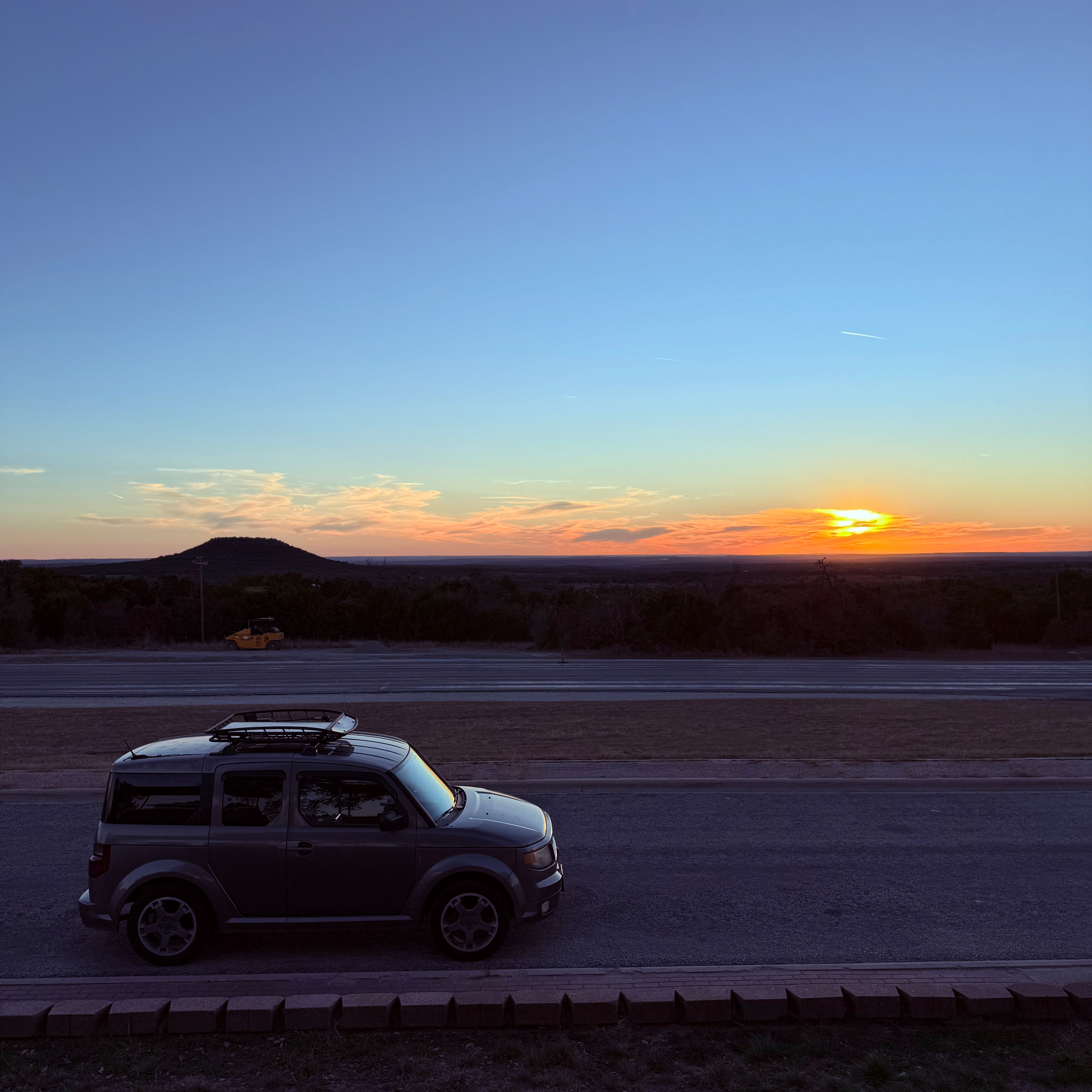 A car is parked on the side of a quiet road with a sunset illuminating the sky and landscape in the background.