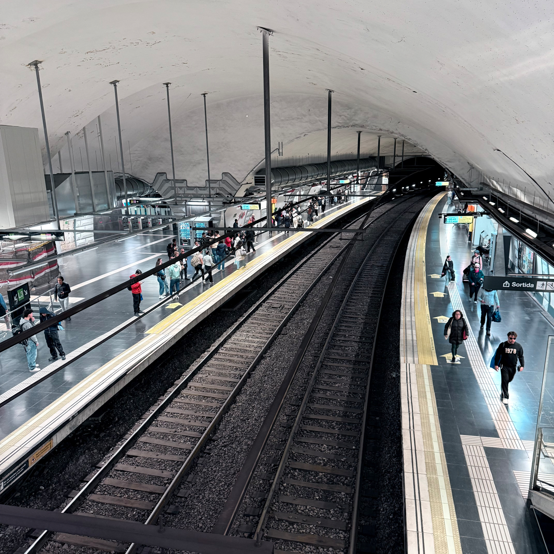 A curved train platform in an underground metro station features people on both sides and tracks in the middle.