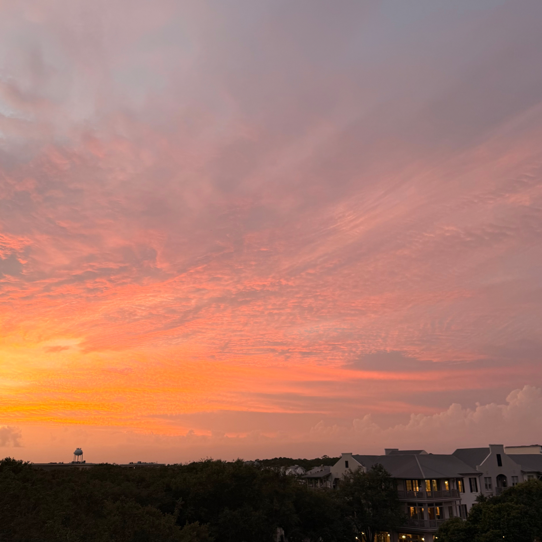 A vibrant sunset paints the sky with shades of orange and pink above a residential area surrounded by trees.