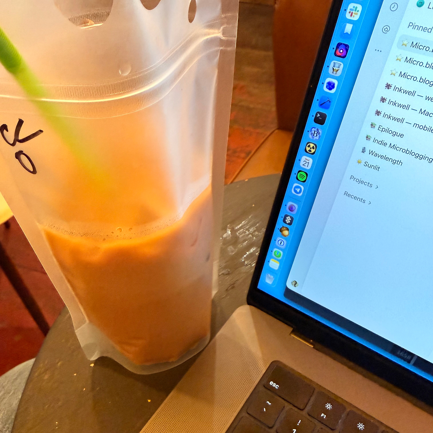 A clear plastic pouch with iced coffee and a green straw is placed next to an open laptop showing Codex with some projects in the sidebar.