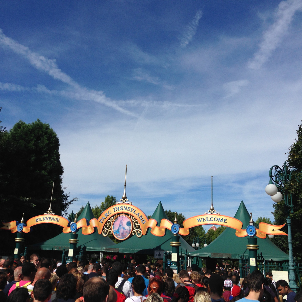 A crowd gathers in front of the entrance to Disneyland Paris, featuring a decorative archway with flags and welcome signs.
