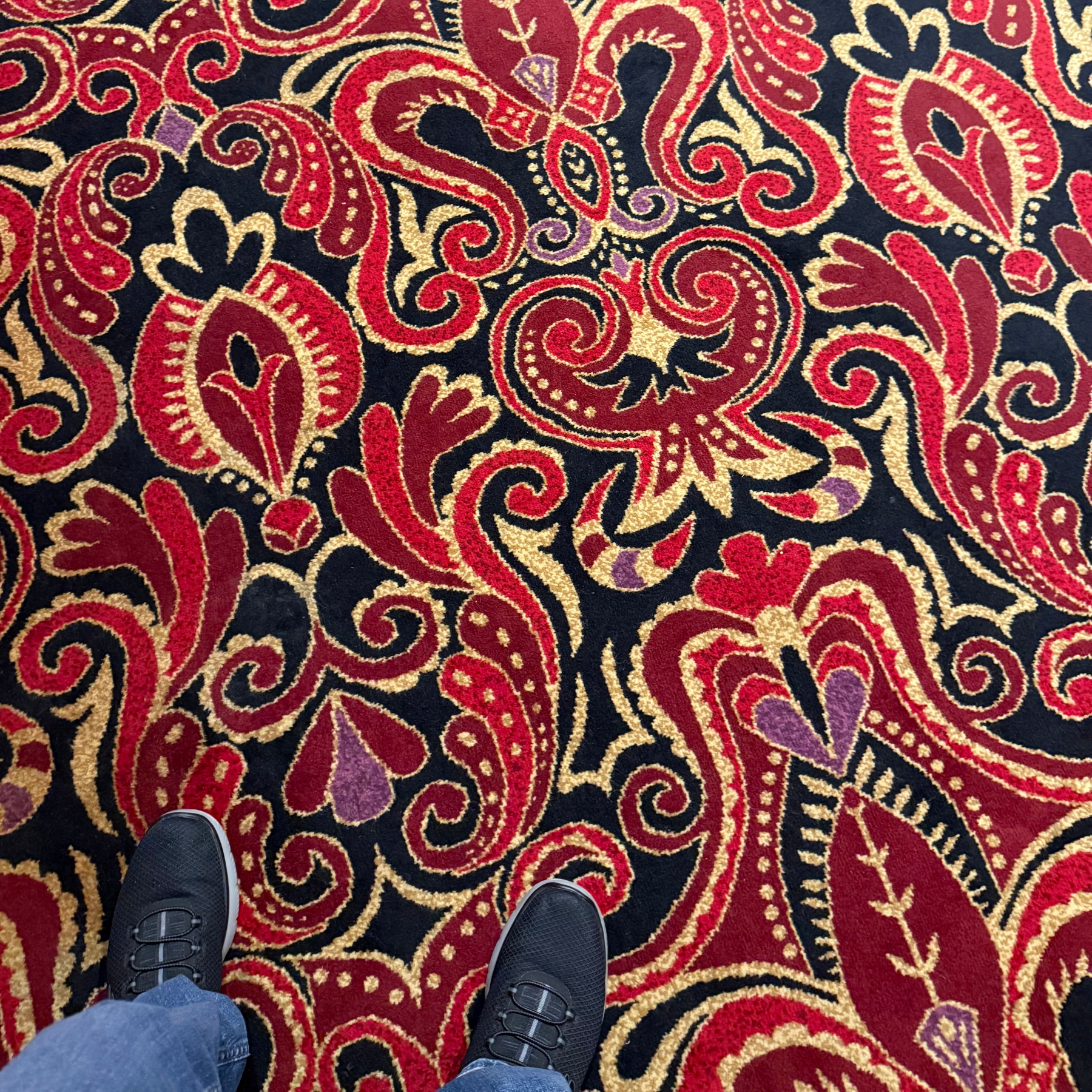 A person stands on a vibrant carpet featuring an ornate pattern with red, yellow, and purple swirls and floral designs.