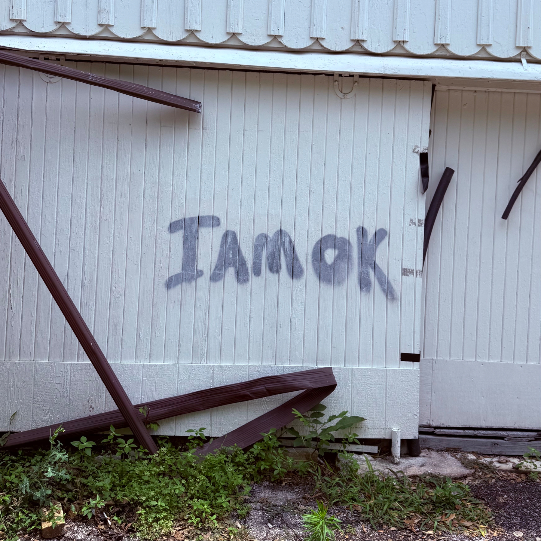 Graffiti reading I AM OK is spray-painted on a weathered, white wooden wall with broken beams leaning against it.