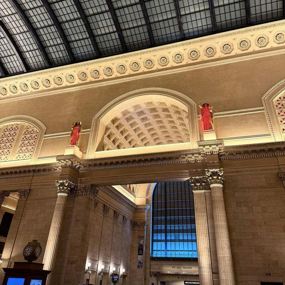 A grand architectural interior features classical columns, two statues draped in red robes, intricate archways, and a glass-paneled ceiling with warm lighting.