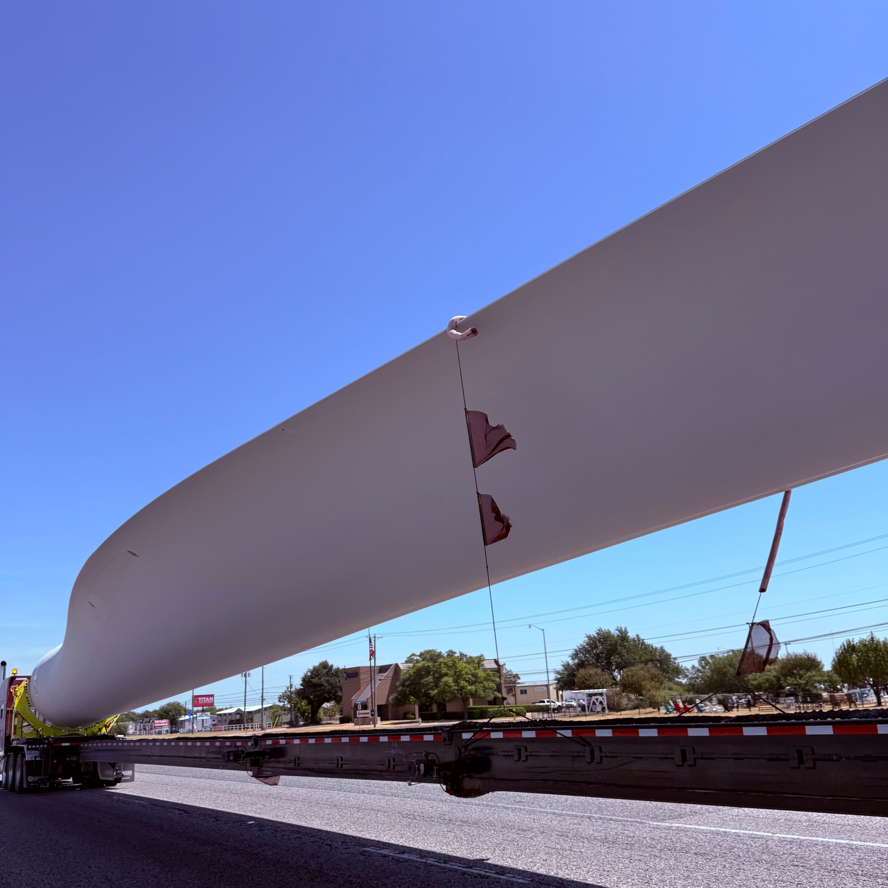 A large wind turbine blade is being transported on a flatbed truck along a road under a clear blue sky.
