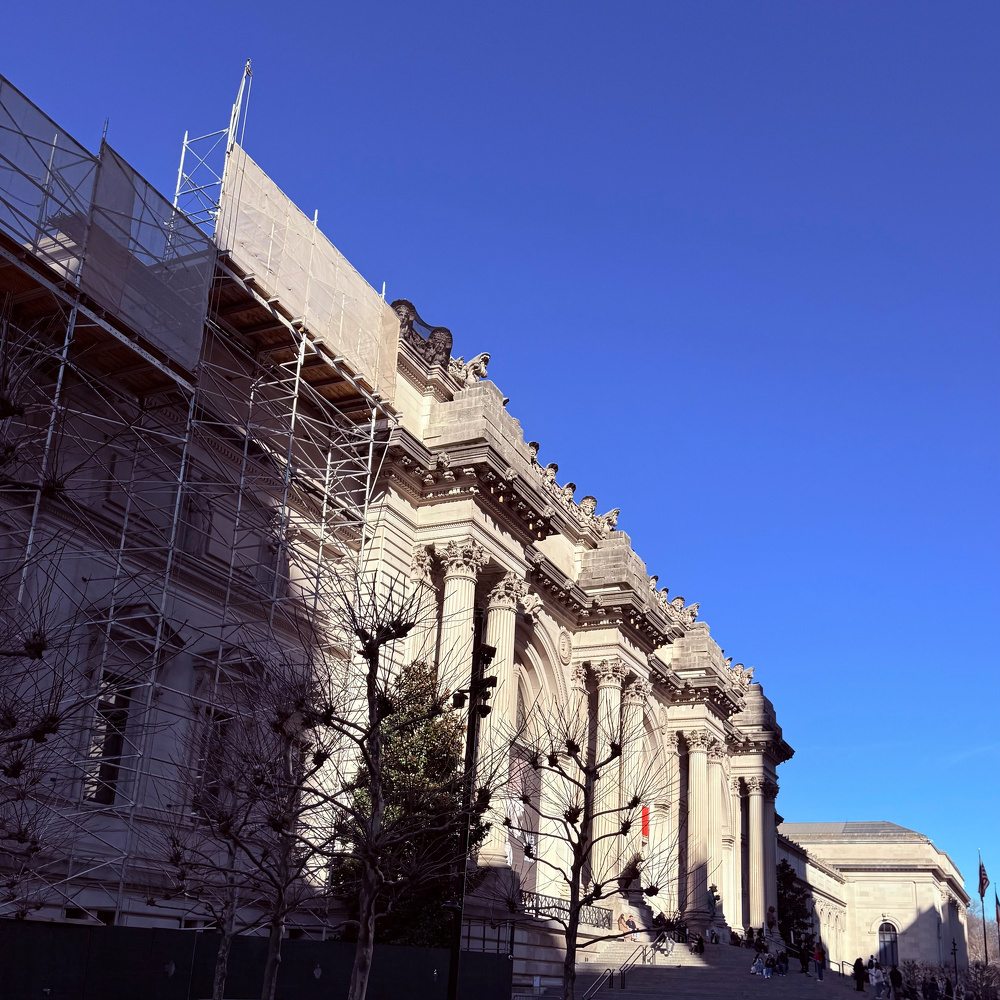 A large neoclassical building with columns is undergoing renovation, with scaffolding on one side and a clear blue sky above.