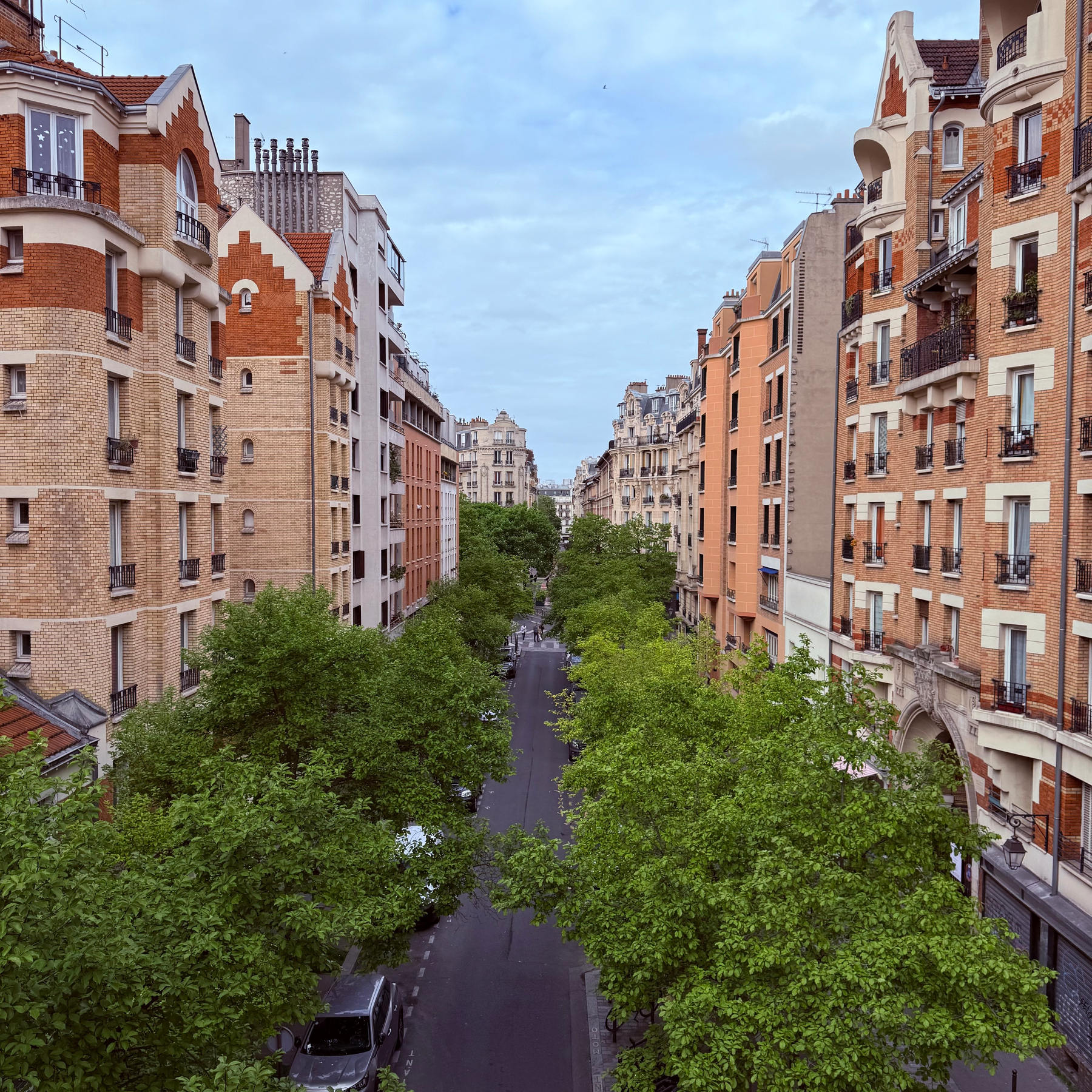A tree-lined street is flanked by charming multi-story buildings with a mix of brick and stucco facades.