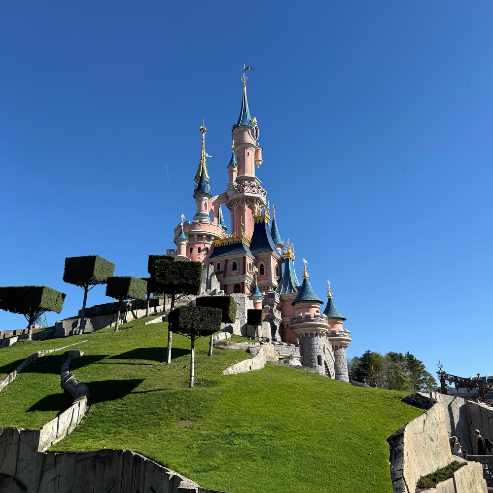 A fairytale castle with pink towers and golden spires sits atop a grassy hill, surrounded by uniquely trimmed trees under a clear blue sky.