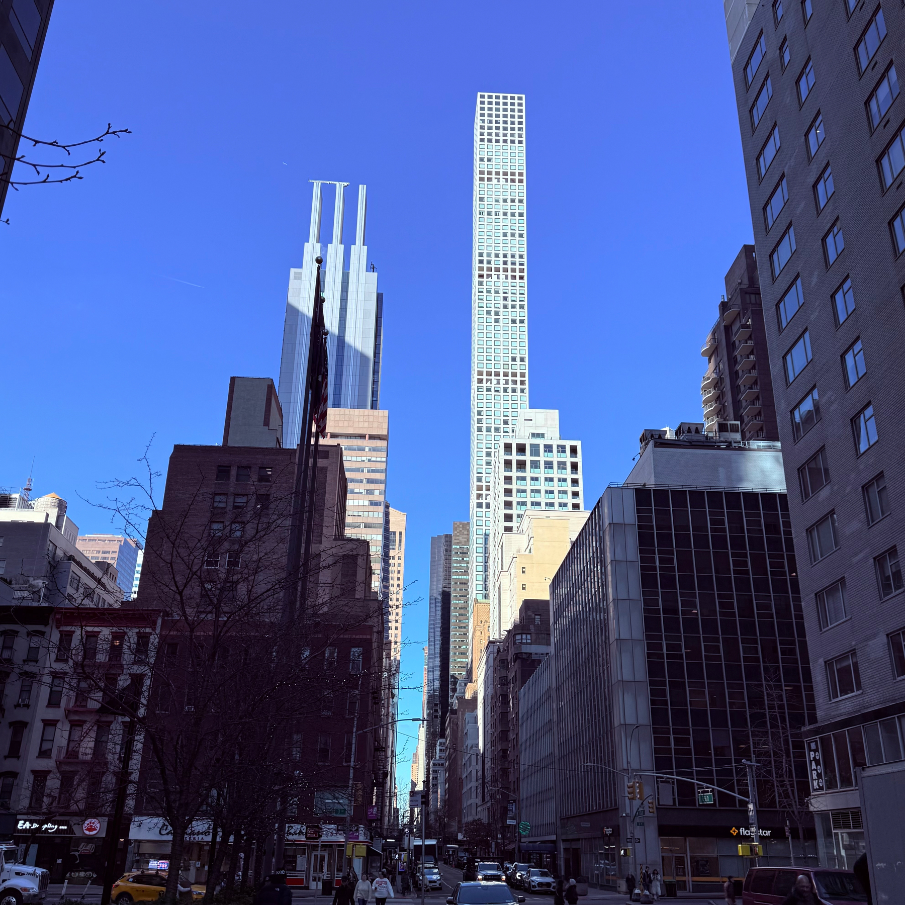 A city street is lined with tall buildings, including a prominent slender skyscraper under a clear blue sky.