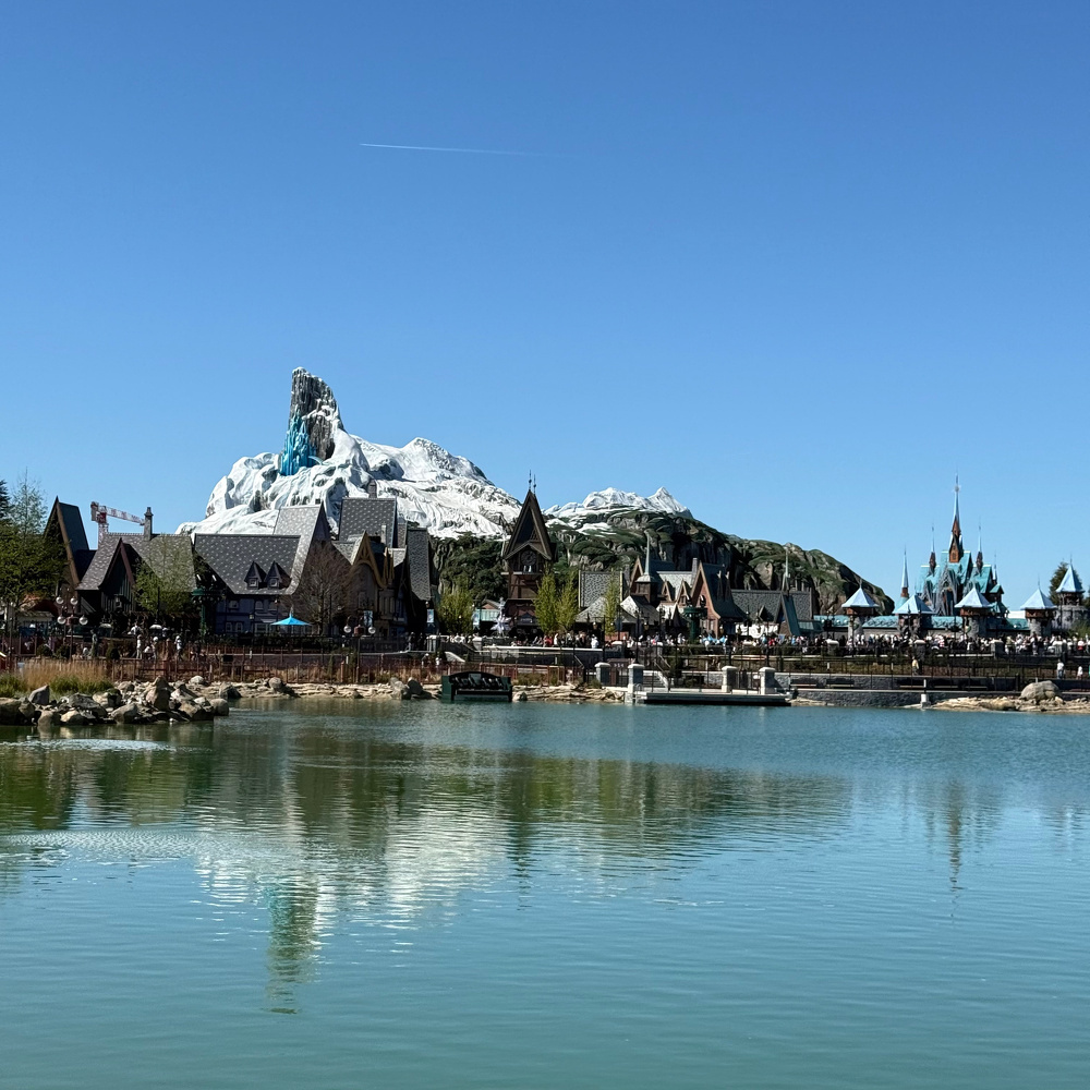 A picturesque theme park scene features a lake, rustic buildings, and a snow-capped mountain in the background.