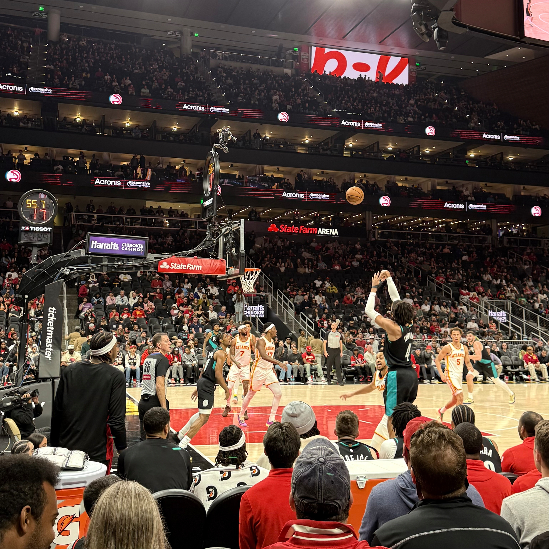 A basketball game is taking place with a player shooting the ball while others on the court and spectators in the arena watch.