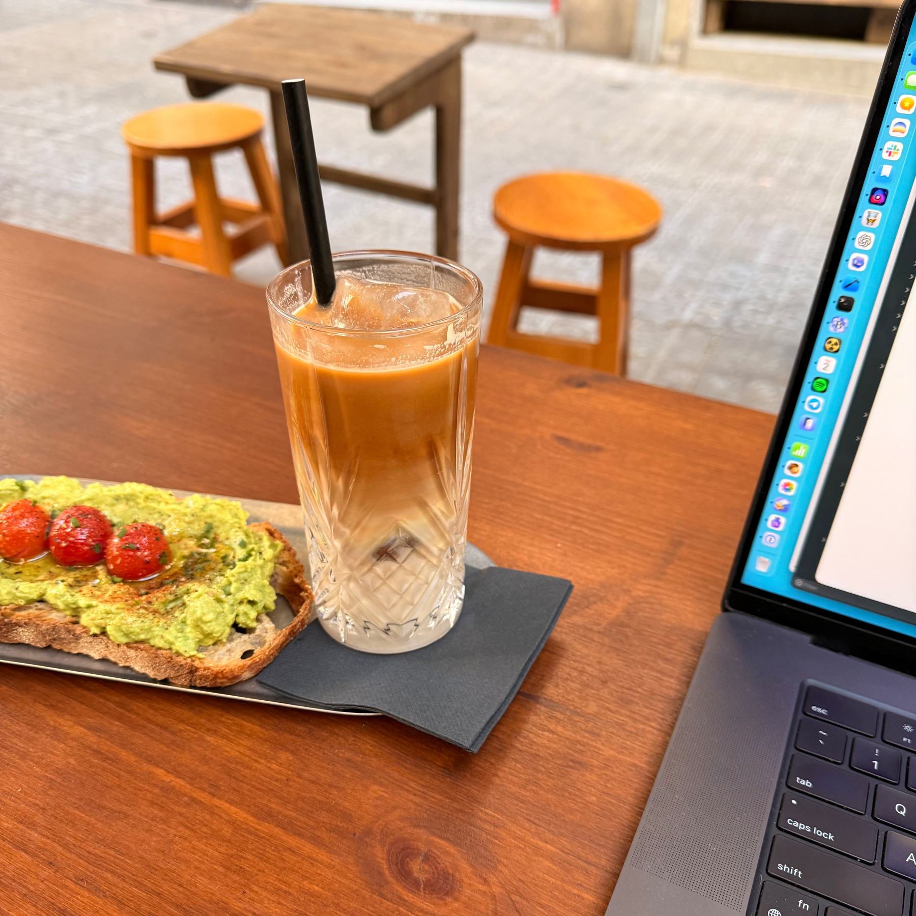 A plate with avocado toast topped with cherry tomatoes, accompanied by an iced coffee, is placed on a wooden table with a laptop and stools in the background.