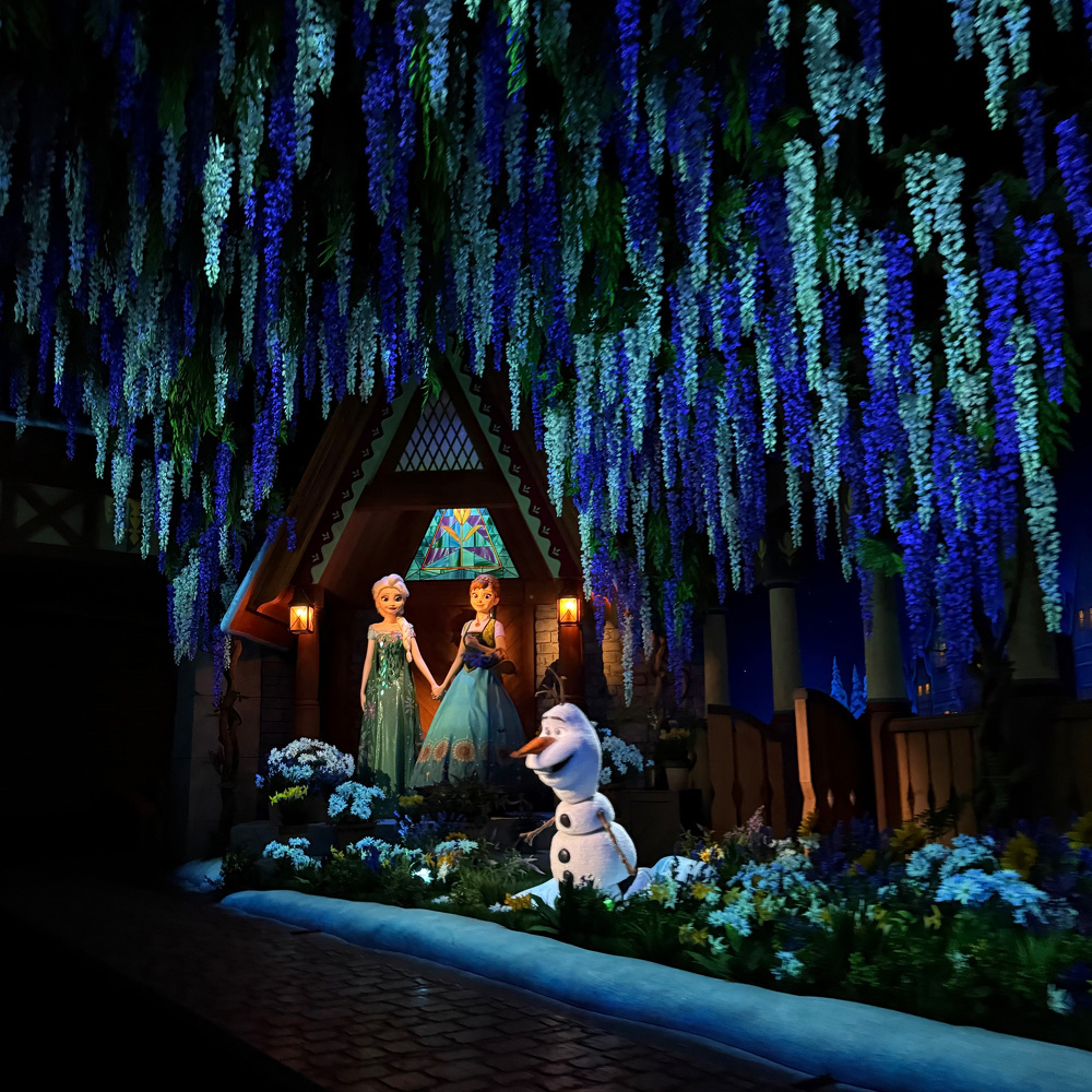 Two animated characters, one in a green dress and another in a blue dress, are standing in a wintry scene with a friendly snowman surrounded by glowing blue and green foliage.