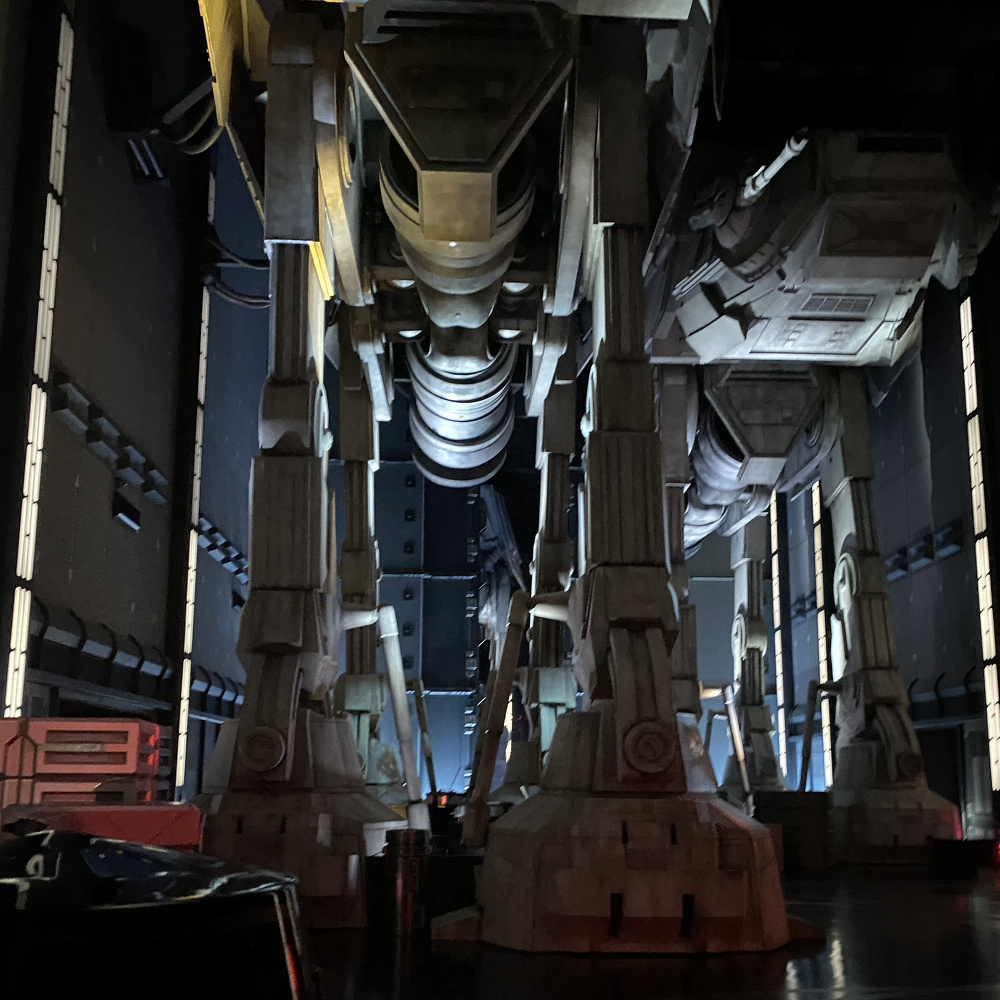 A futuristic, industrial scene showcases the legs of two massive mechanical walkers in a dimly lit environment.
