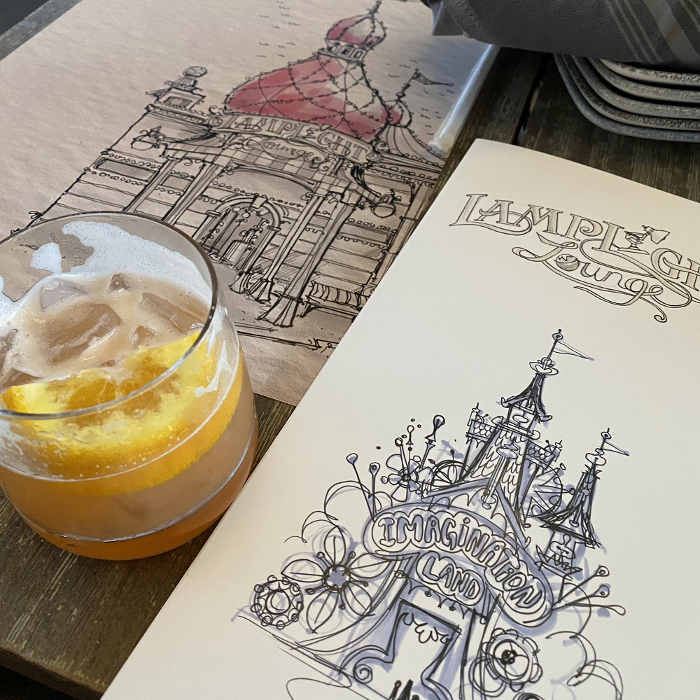 A cocktail with an orange garnish sits on a table alongside two sketches featuring architectural drawings labeled Lamplight Lounge and Imagination Land.