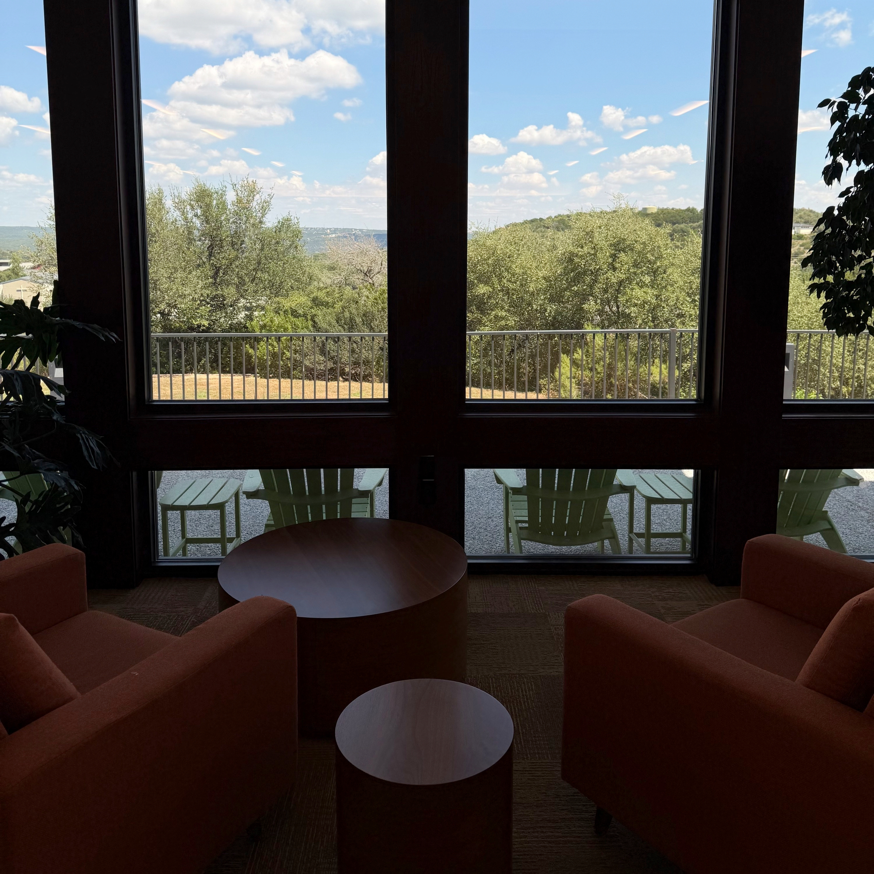 A cozy indoor seating area with two armchairs and a round table overlooks a balcony with Adirondack chairs and a scenic view of trees and hills under a partly cloudy sky.