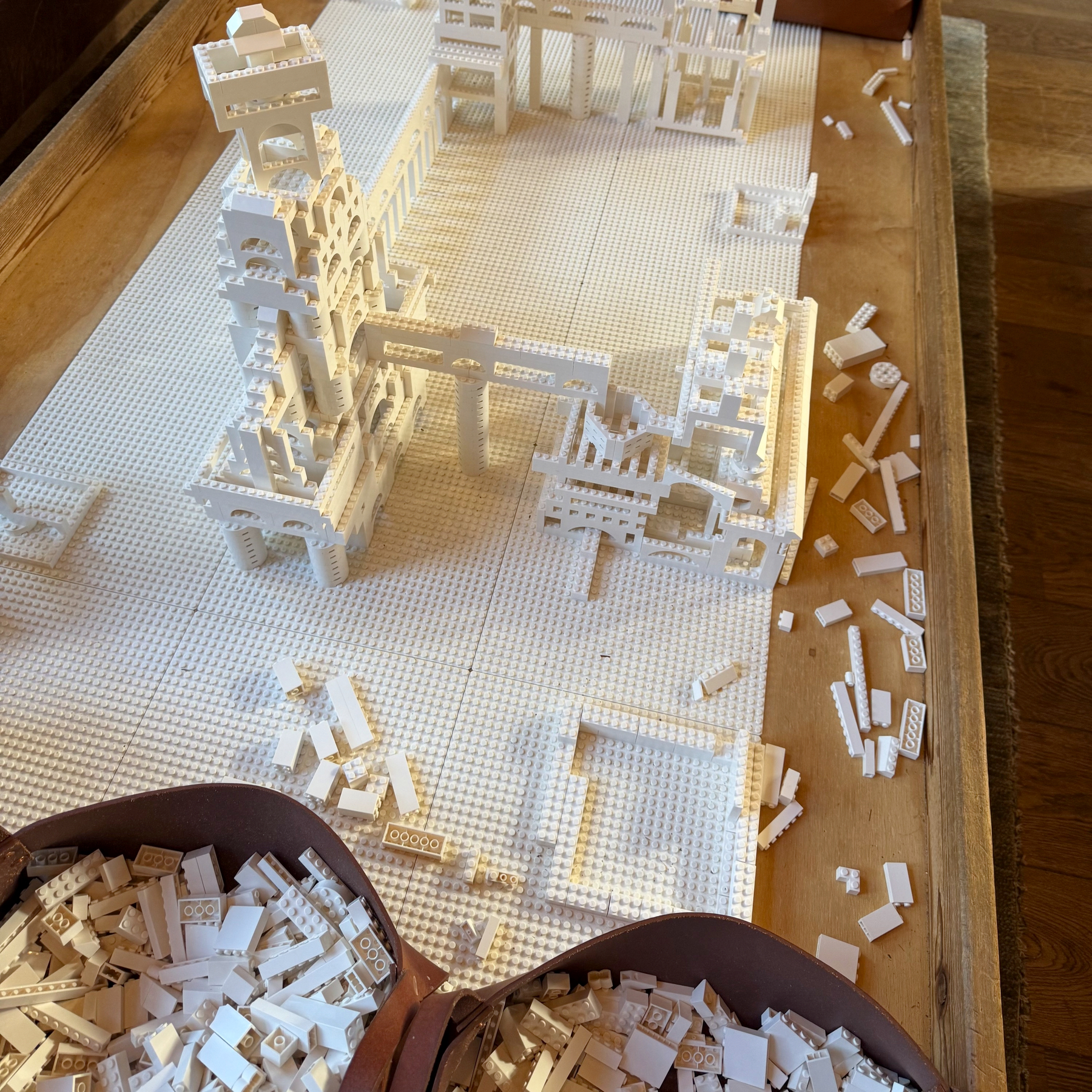 A partially constructed white LEGO structure resembling architectural buildings is placed on a large baseplate, with numerous small white LEGO pieces scattered around and stored in containers nearby.