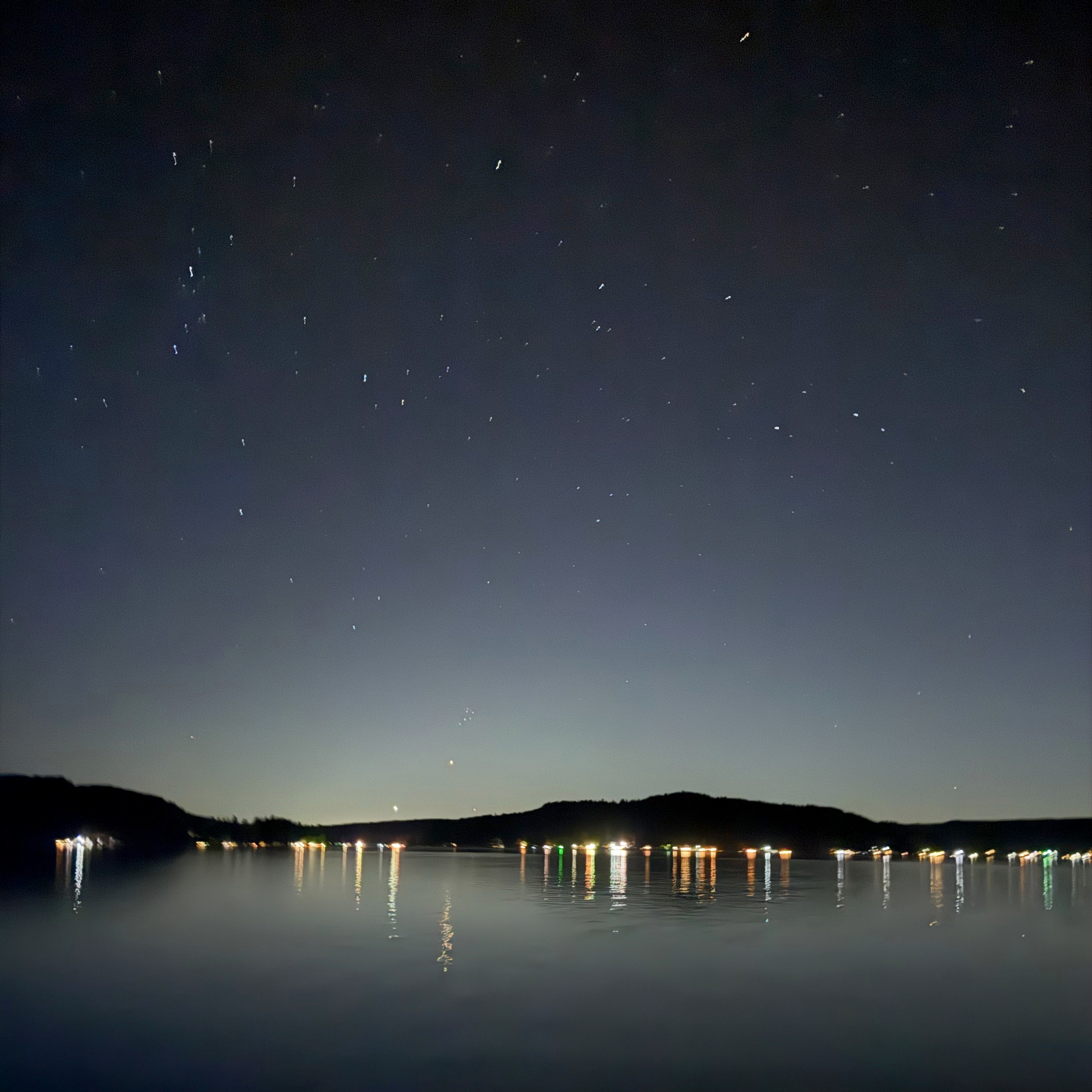 A calm lake at night with reflections of lights from a distant shore and a starry sky above.