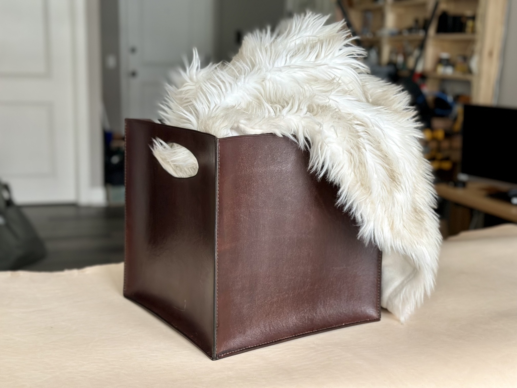 A rather beautiful chocolate dyed leather bin sitting sitting on a table with a large and fluffy white blanket protruding from the top. The bin itself is 12 inches square and has handles cut out on two sides for easy grabbing.