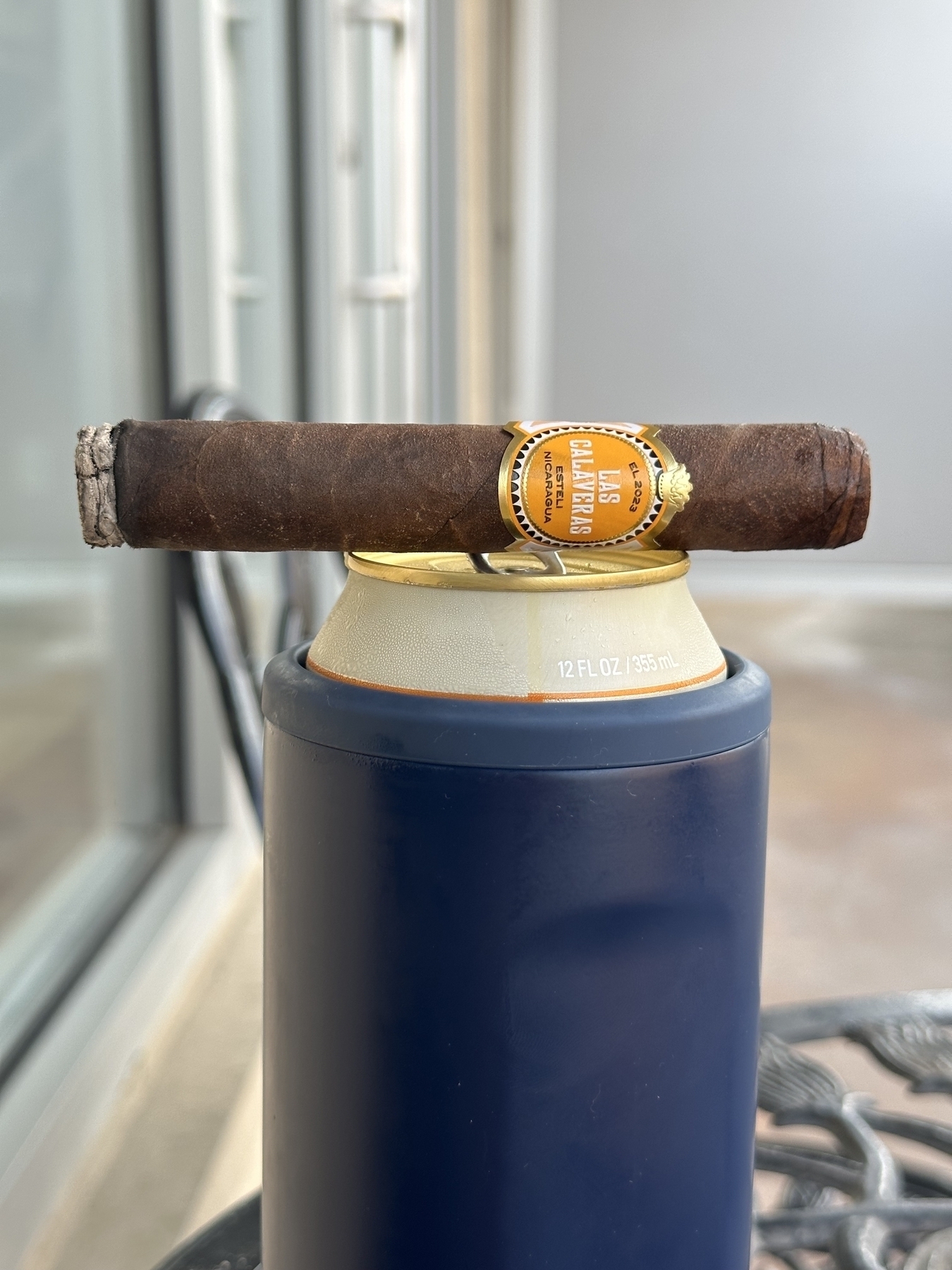 A lit Las Calaveras EL 2023 cigar on top of an open beer can which is inside of a blue insulated beer cozy thingy. 