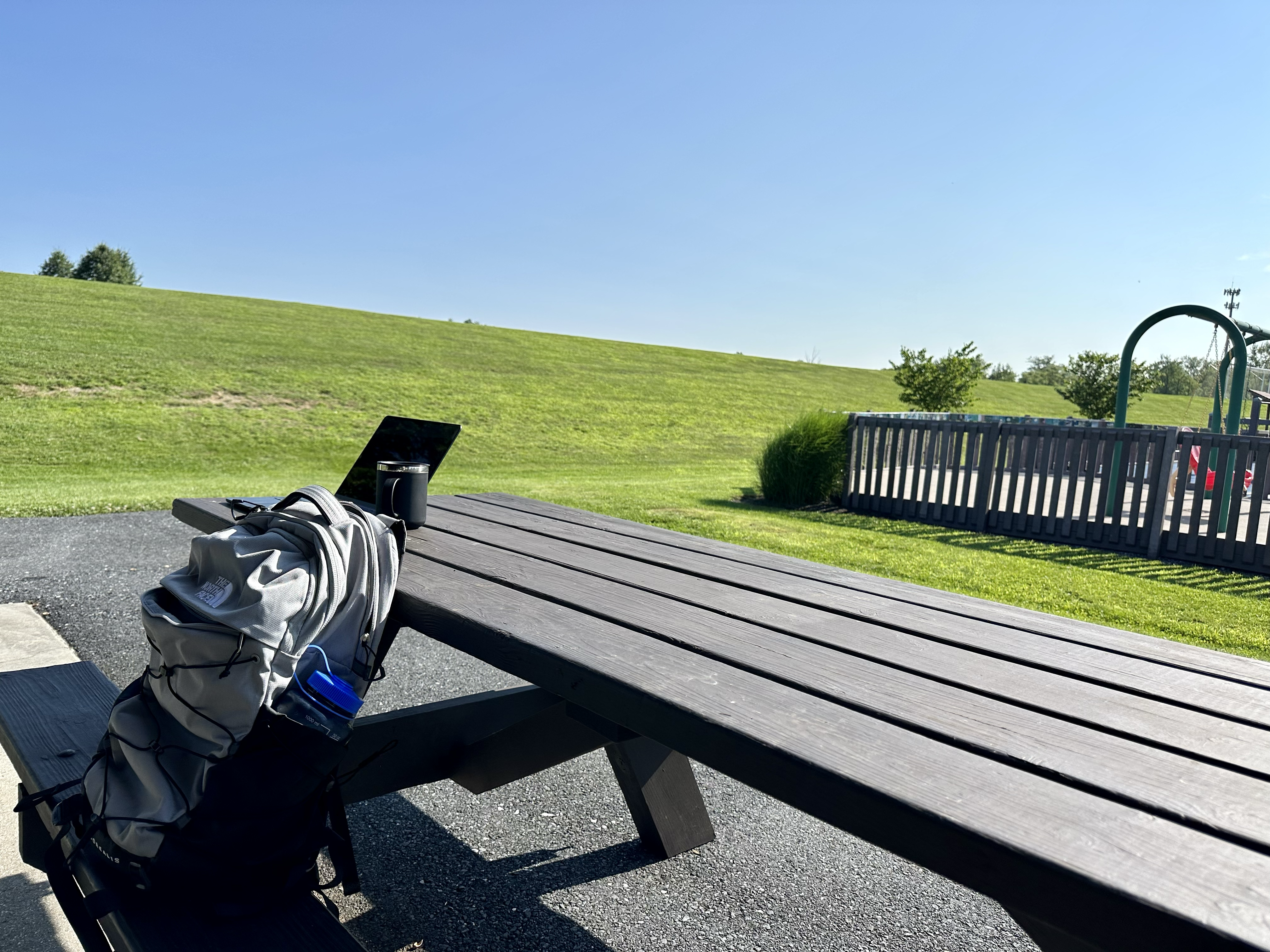 macbook air, coffee mug, and north face back pack resting on a park picnic bench, overlooking a playground on the right side of the photo and a vast green field and blue sky to the left and above.