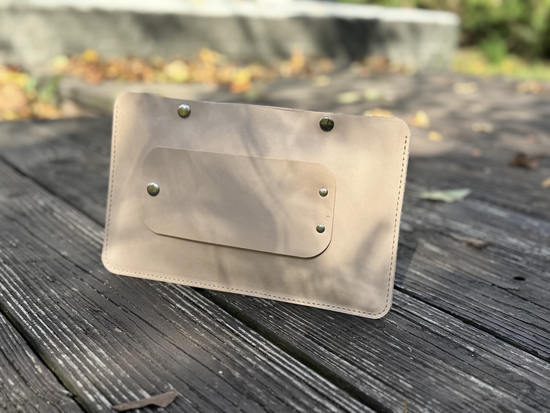 A beige leather wallet with metal studs is placed on a wooden surface outdoors.