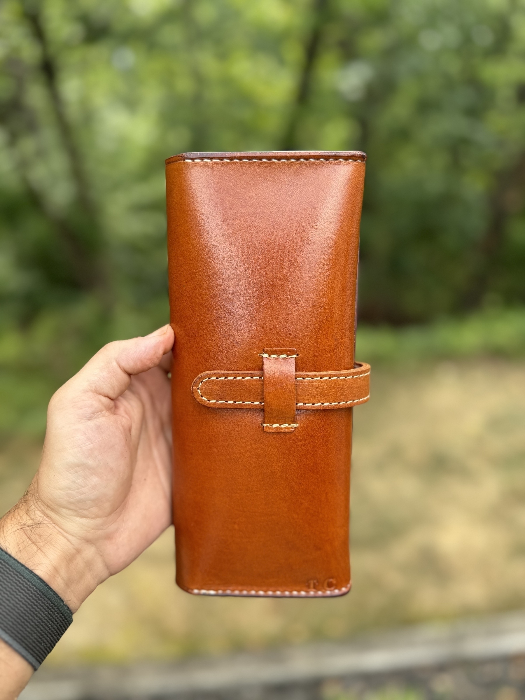 A hand is holding a brown leather cigar case against a blurred outdoor background of greenery and grass.