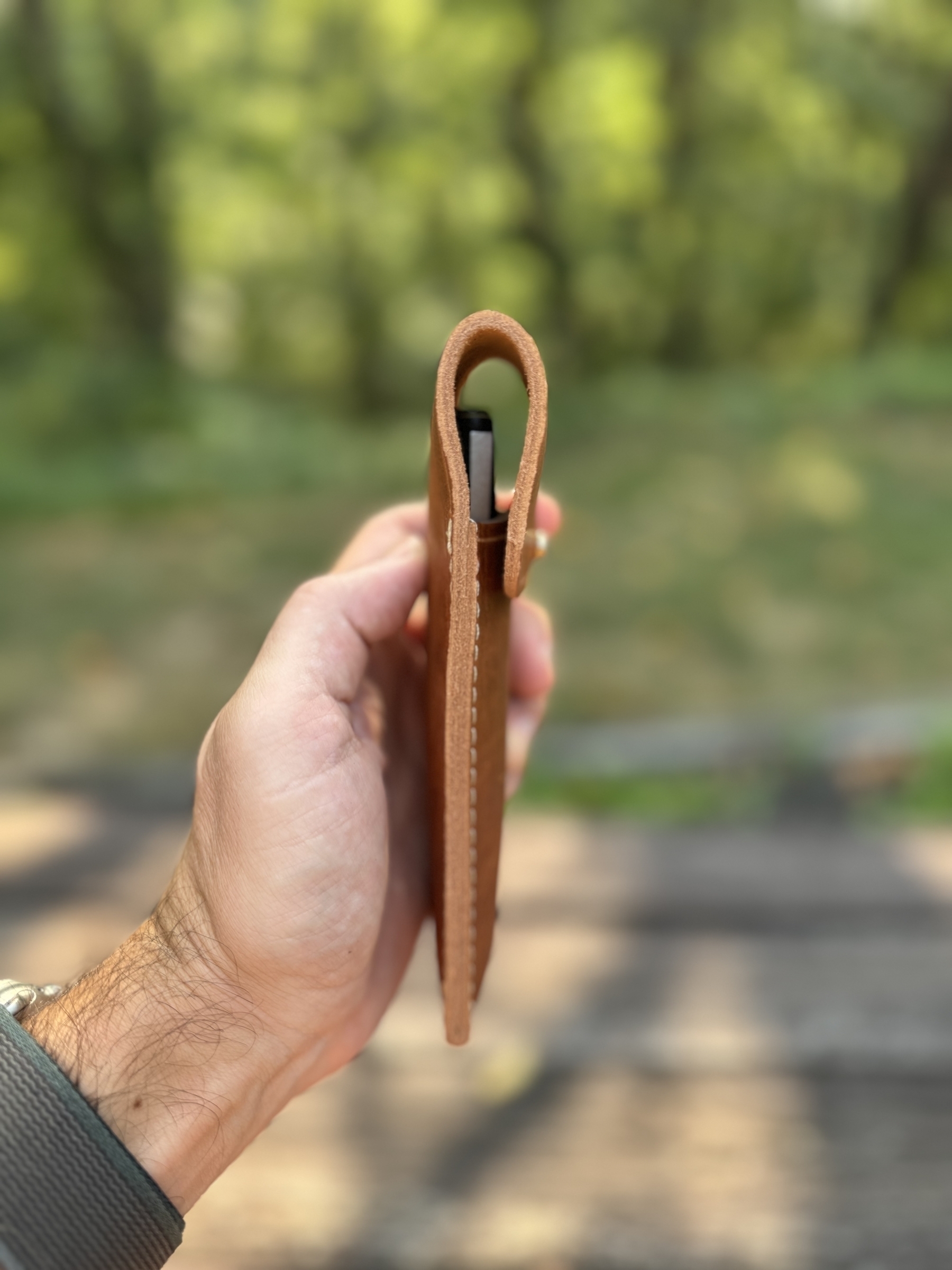 A hand is holding a leather case containing a pen against a blurred outdoor background.