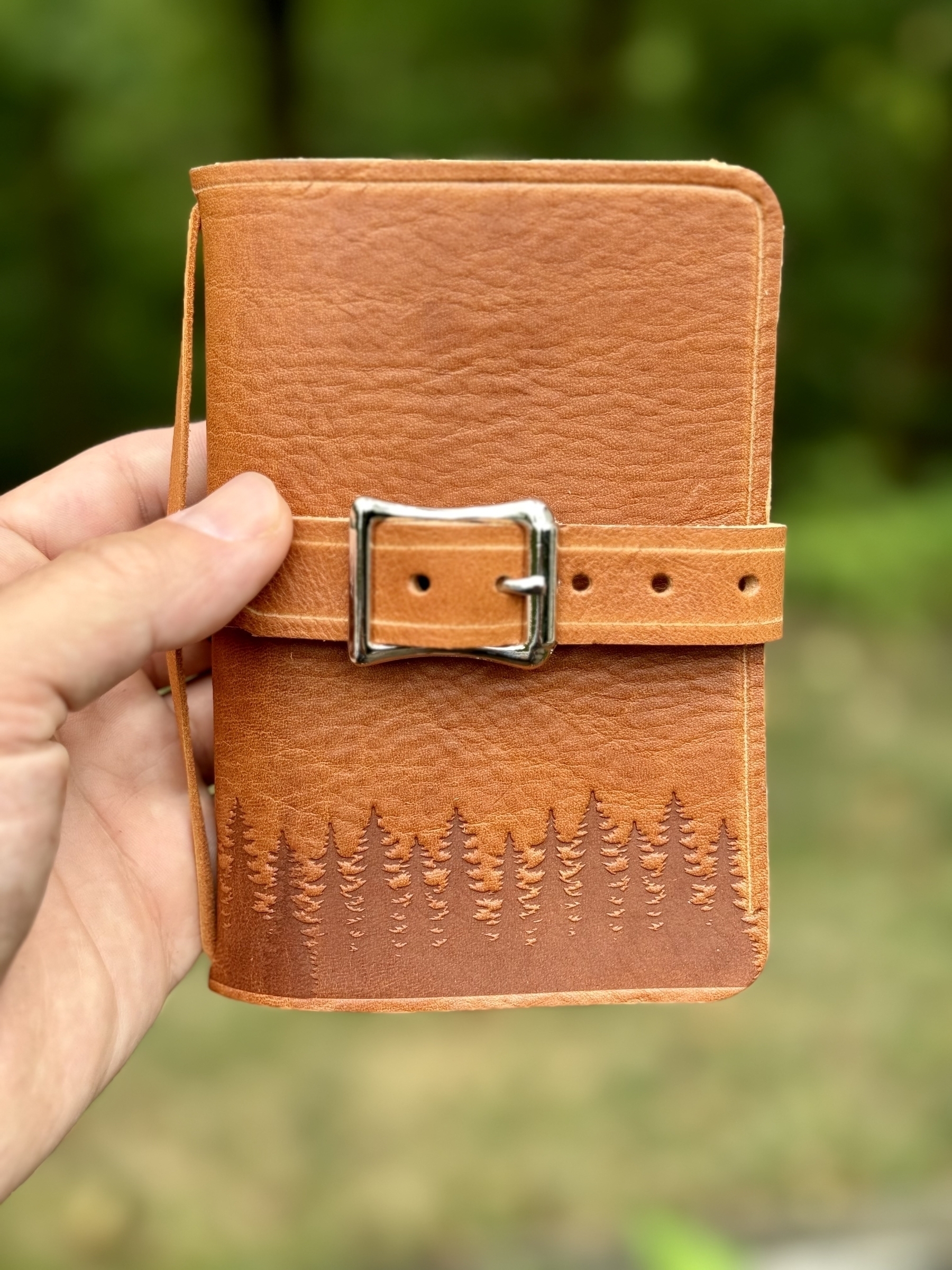 A hand is holding a small tan leather-bound notebook with a metal buckle and an embossed tree design.