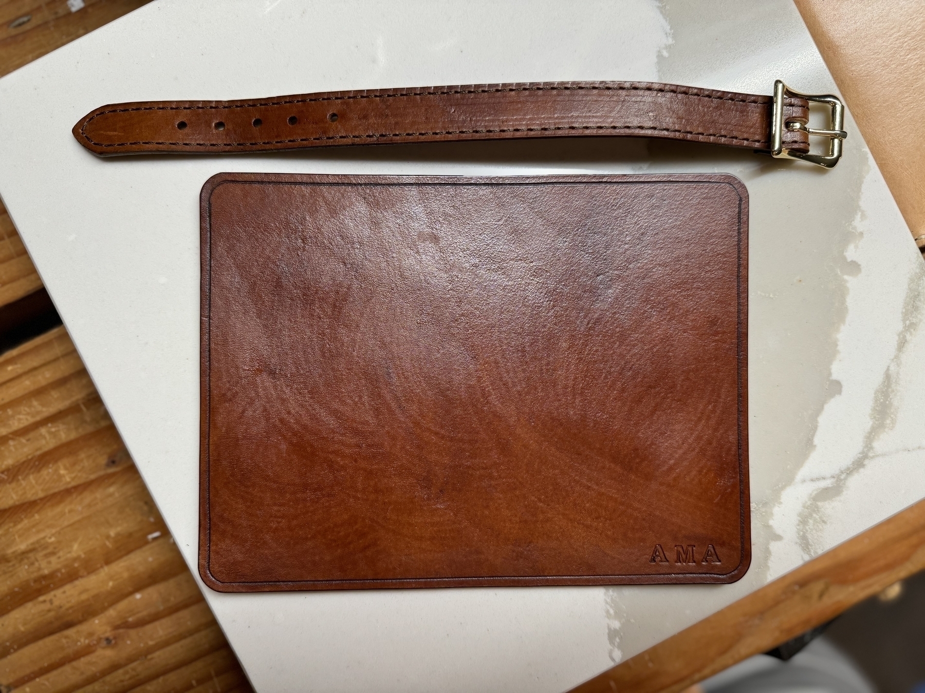 A brown leather strap and a rectangular brown leather object, possibly a wallet or a case, are placed on a wooden and white surface.