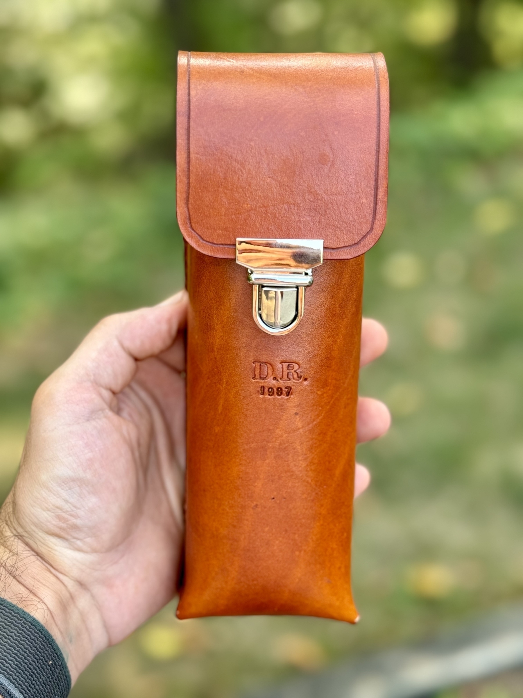 A hand is holding a brown leather case with a metallic clasp and the initials "D.R." and the year "1987" embossed on it.