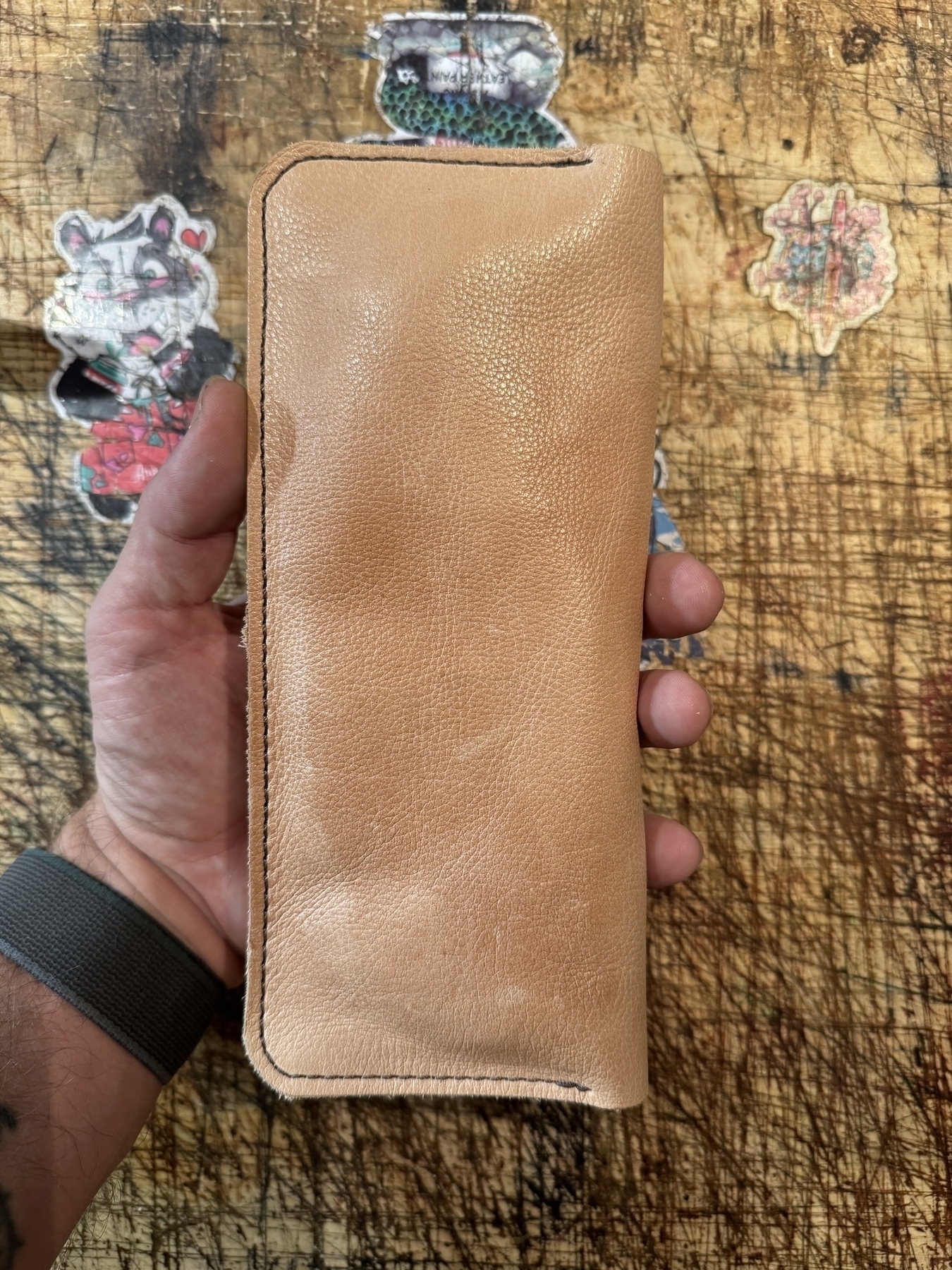 A hand is holding a tan leather wallet above a scratch-covered wooden surface adorned with various stickers.