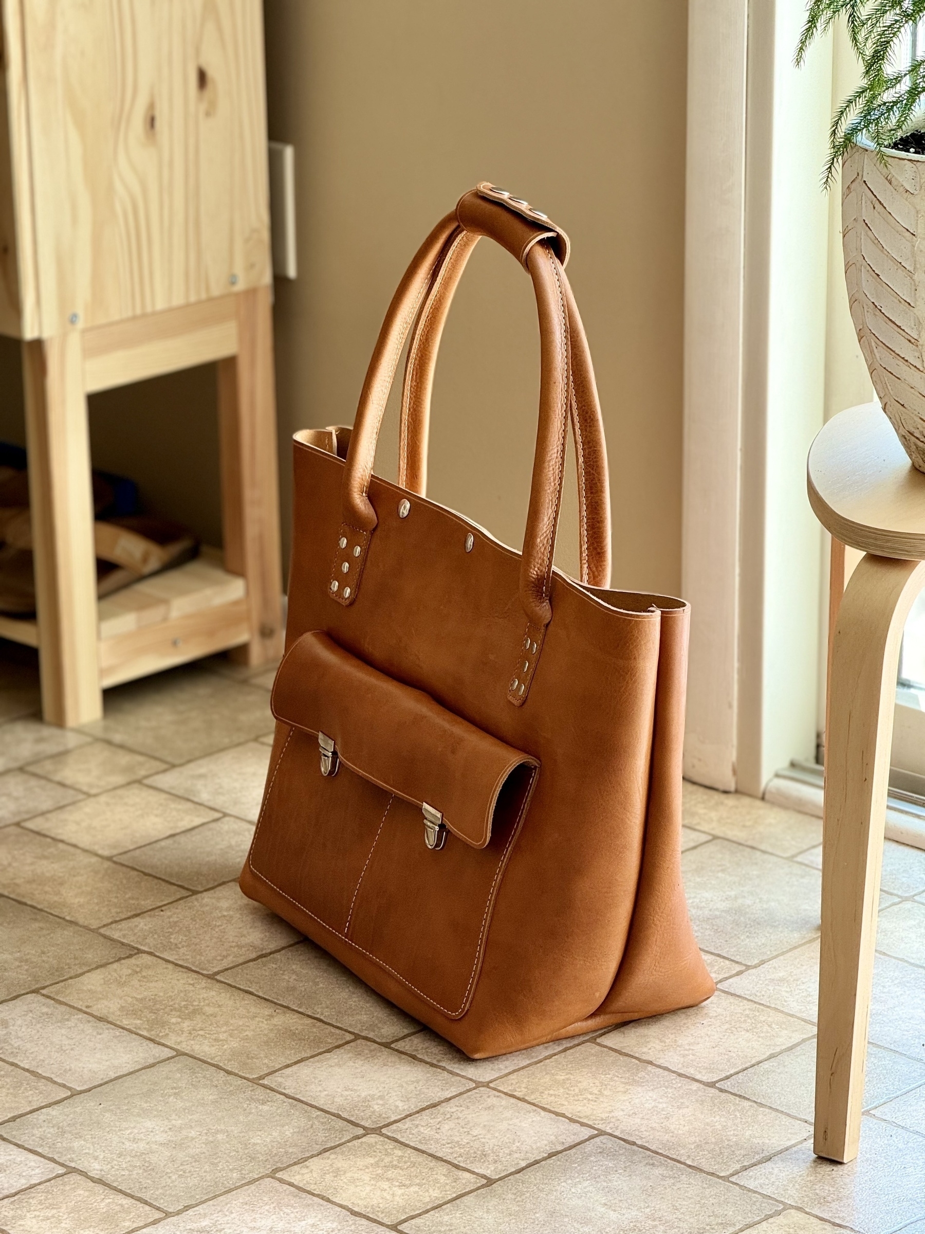 A stylish tan leather handbag (called “Ocean Front”) with a front pocket is placed on a tiled floor near some light-colored wooden furniture.
