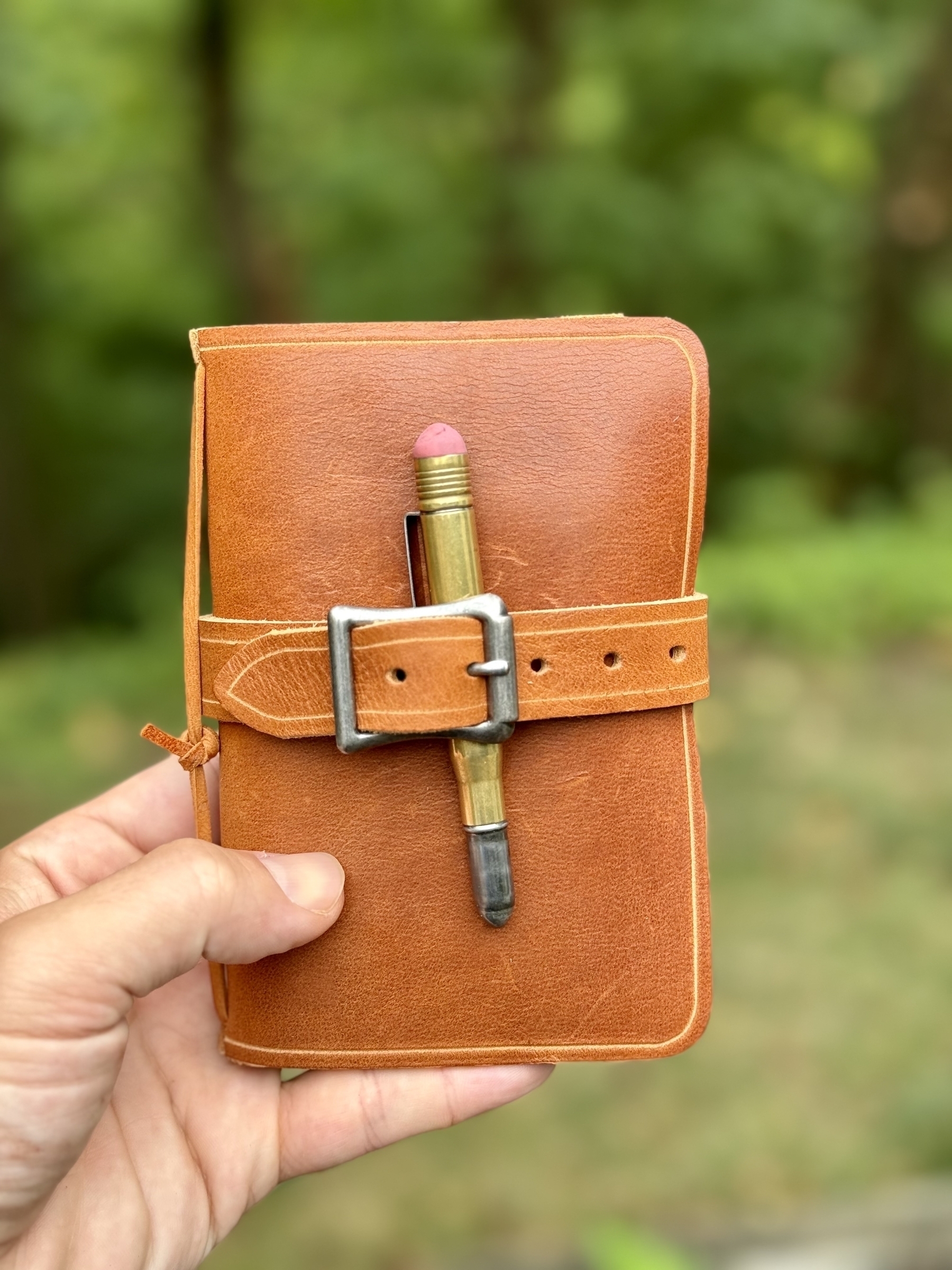 A hand is holding a small tan leather-bound notebook held together by a strap with a pencil placed inside the strap, against a blurred green background.