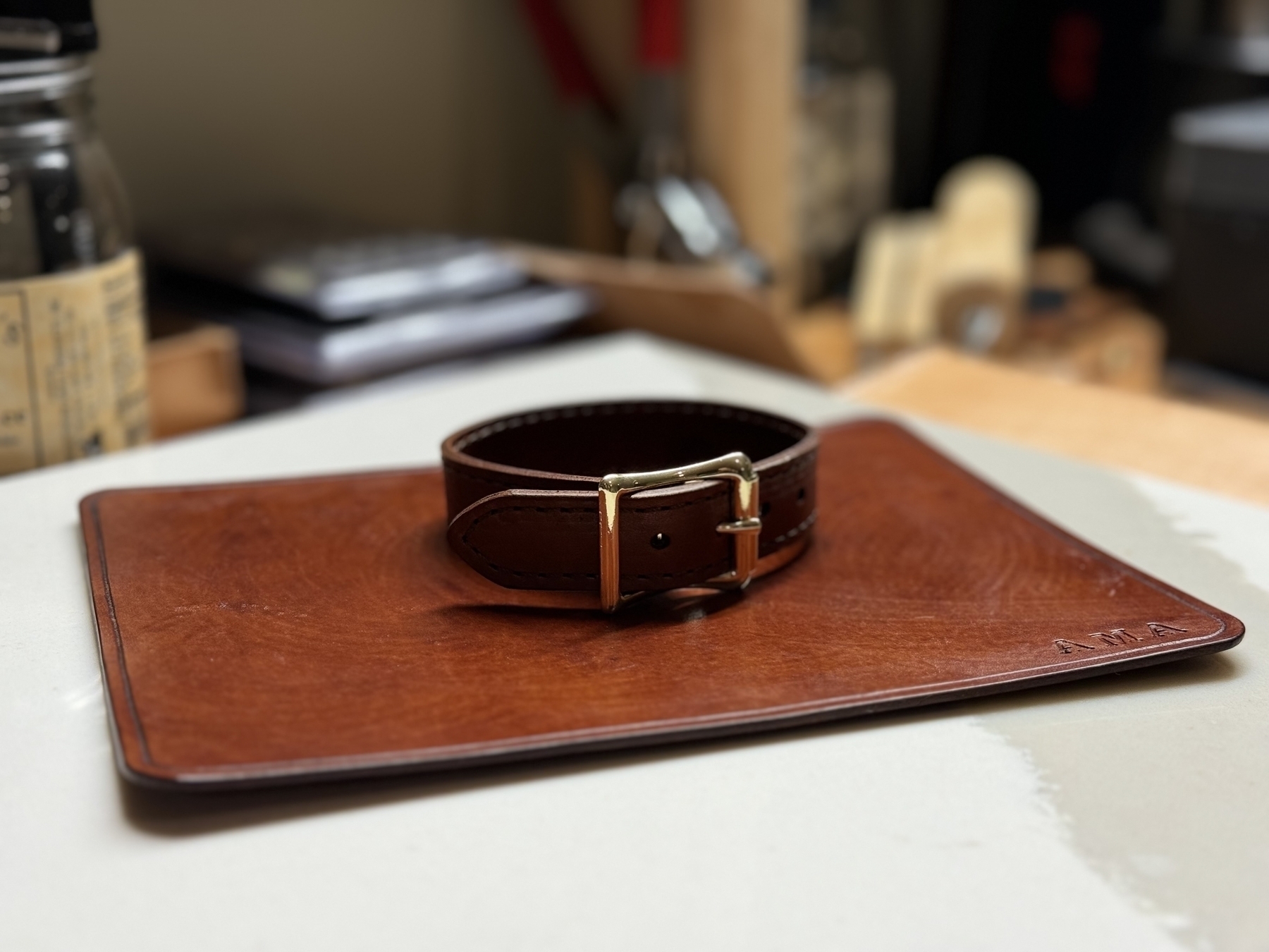 A brown leather strap with a golden buckle is placed on a leather pad on a desk.