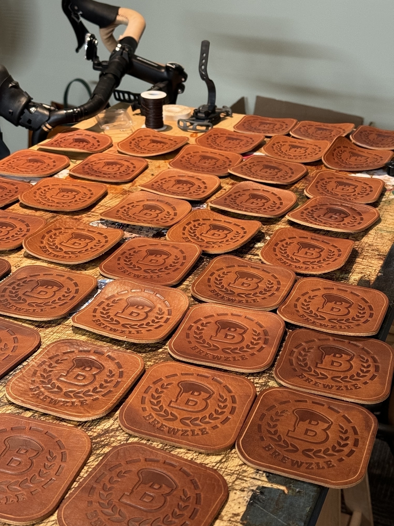 A workbench is covered with numerous brown leather coasters, each embossed with a circular emblem featuring a "B" in the center and surrounded by laurel wreaths.