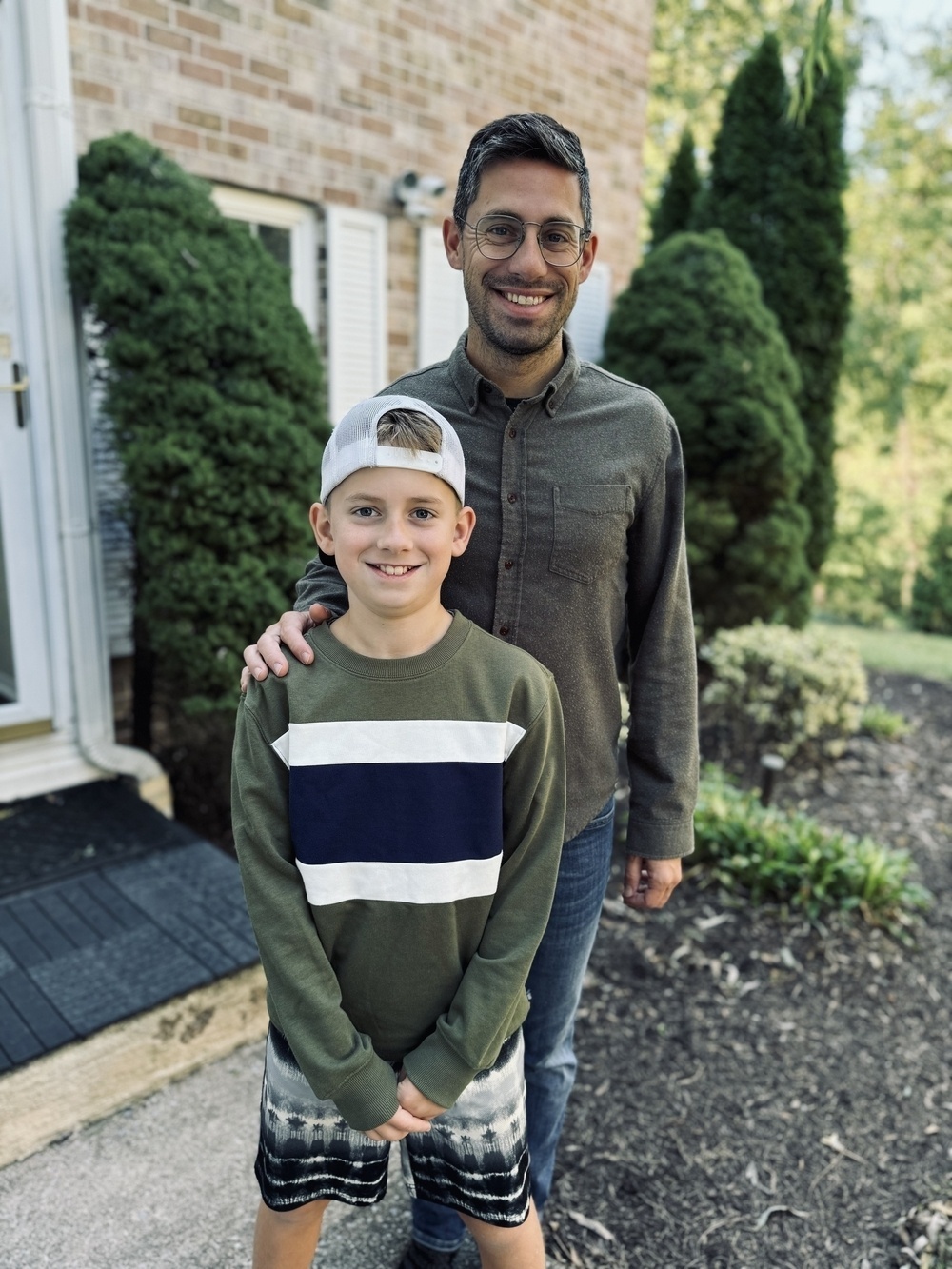 A smiling man stands next to a young boy wearing a cap, posing happily outside a building with bushes in the background.