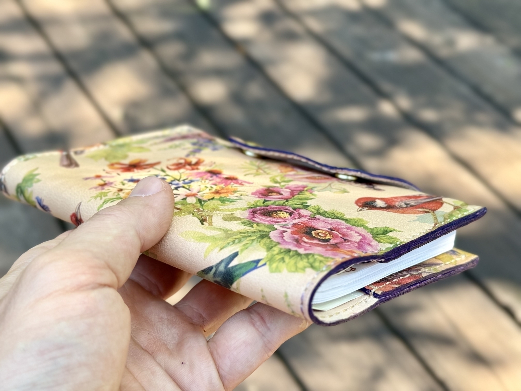 A hand is holding a small book or notebook with a floral and bird design on its cover.