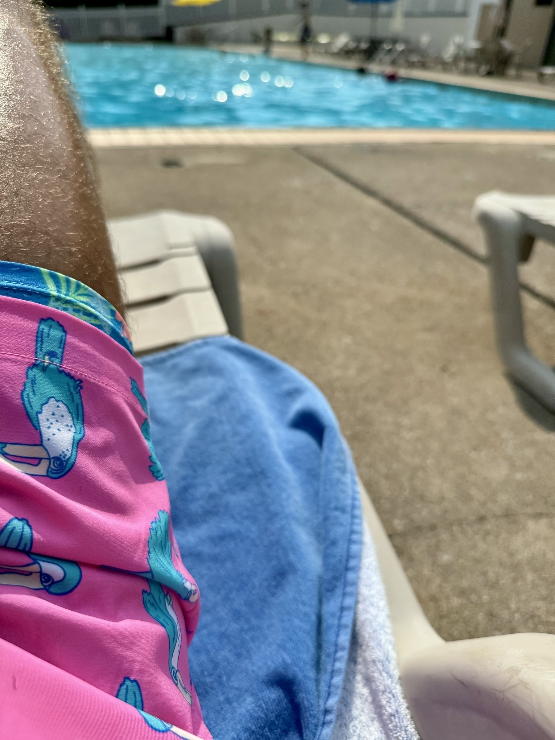 A person with pink swim trunks featuring a swimming bird design is lying on a poolside chair near a swimming pool.