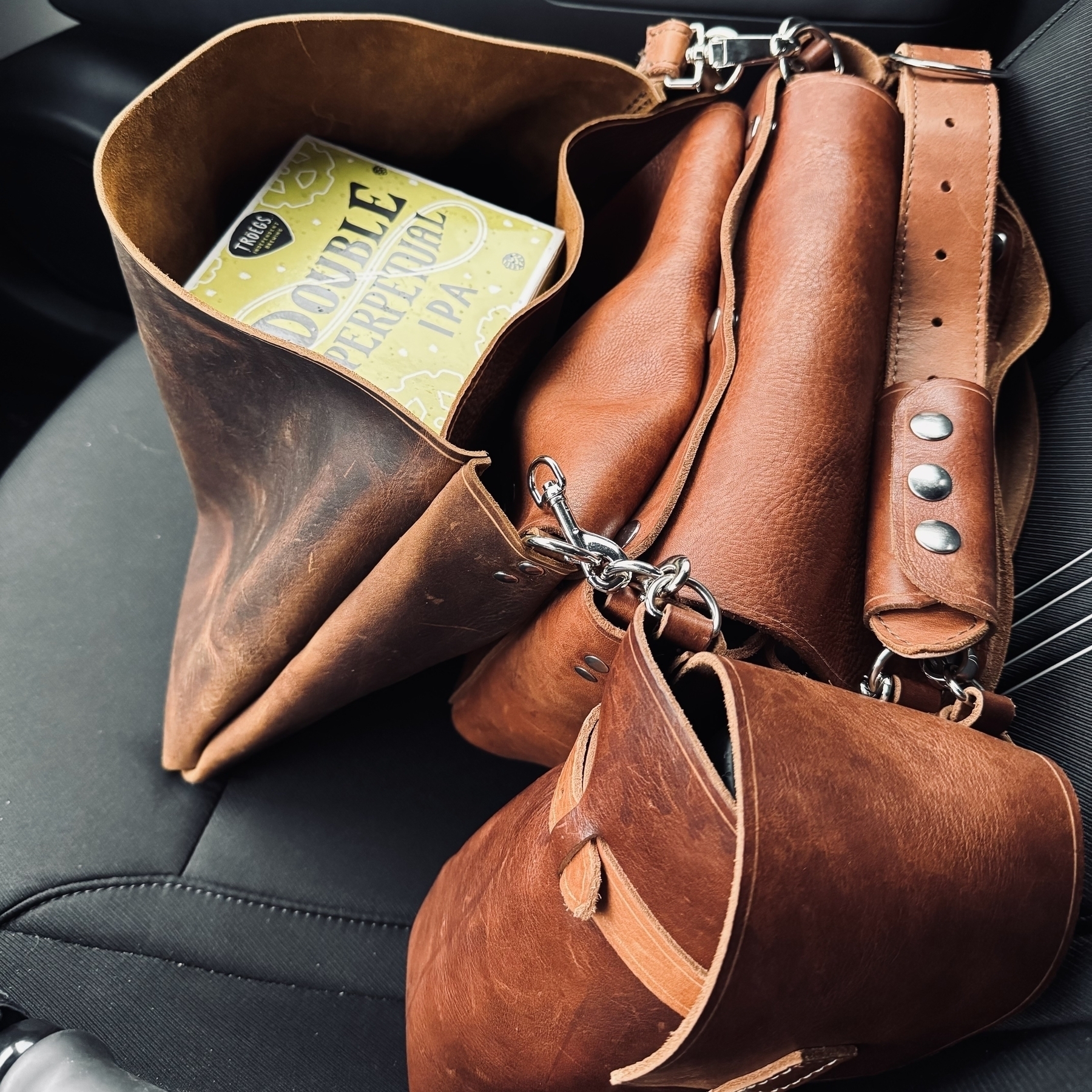 A brown leather bag sits on a car seat, partially open with a can labeled "Double Perceptual IPA" inside.