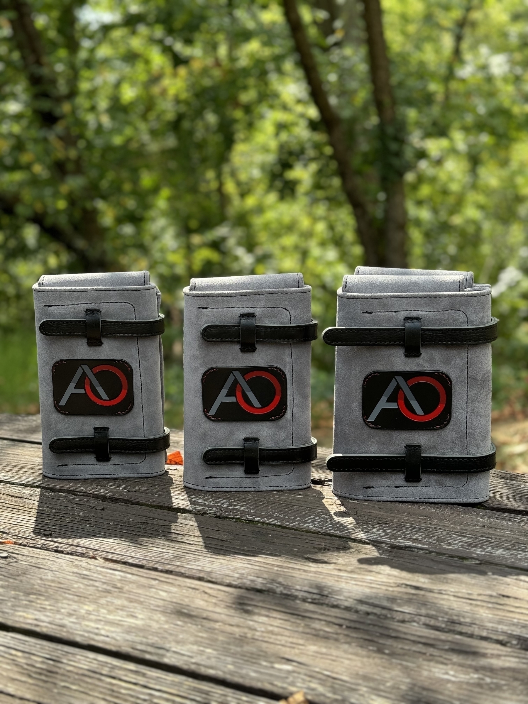 Three gray suede cigar cases with black straps and AO logos are standing on a wooden surface with a forested background.