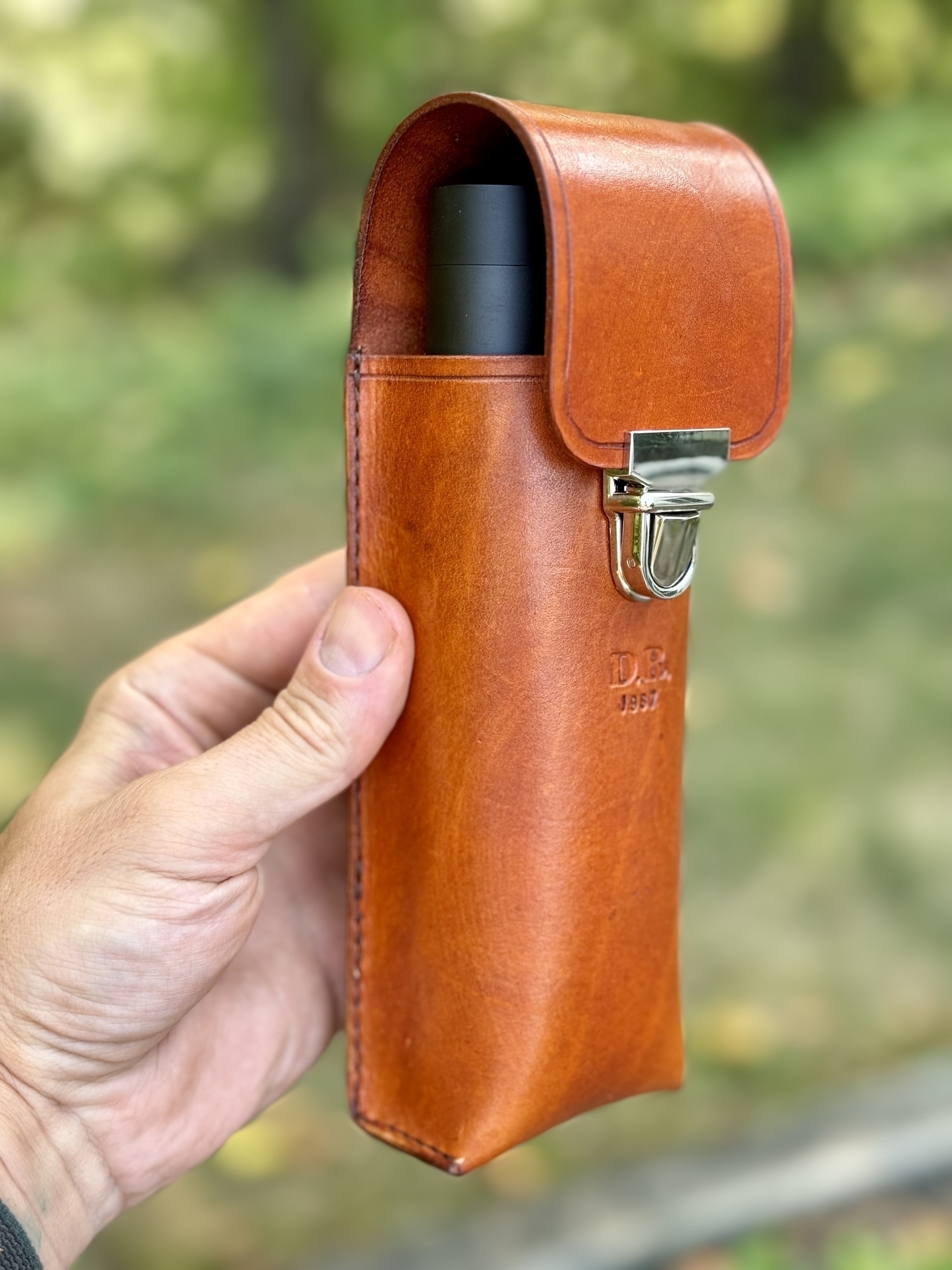 A hand is holding a brown leather case with a top flap, containing a black cylindrical object.