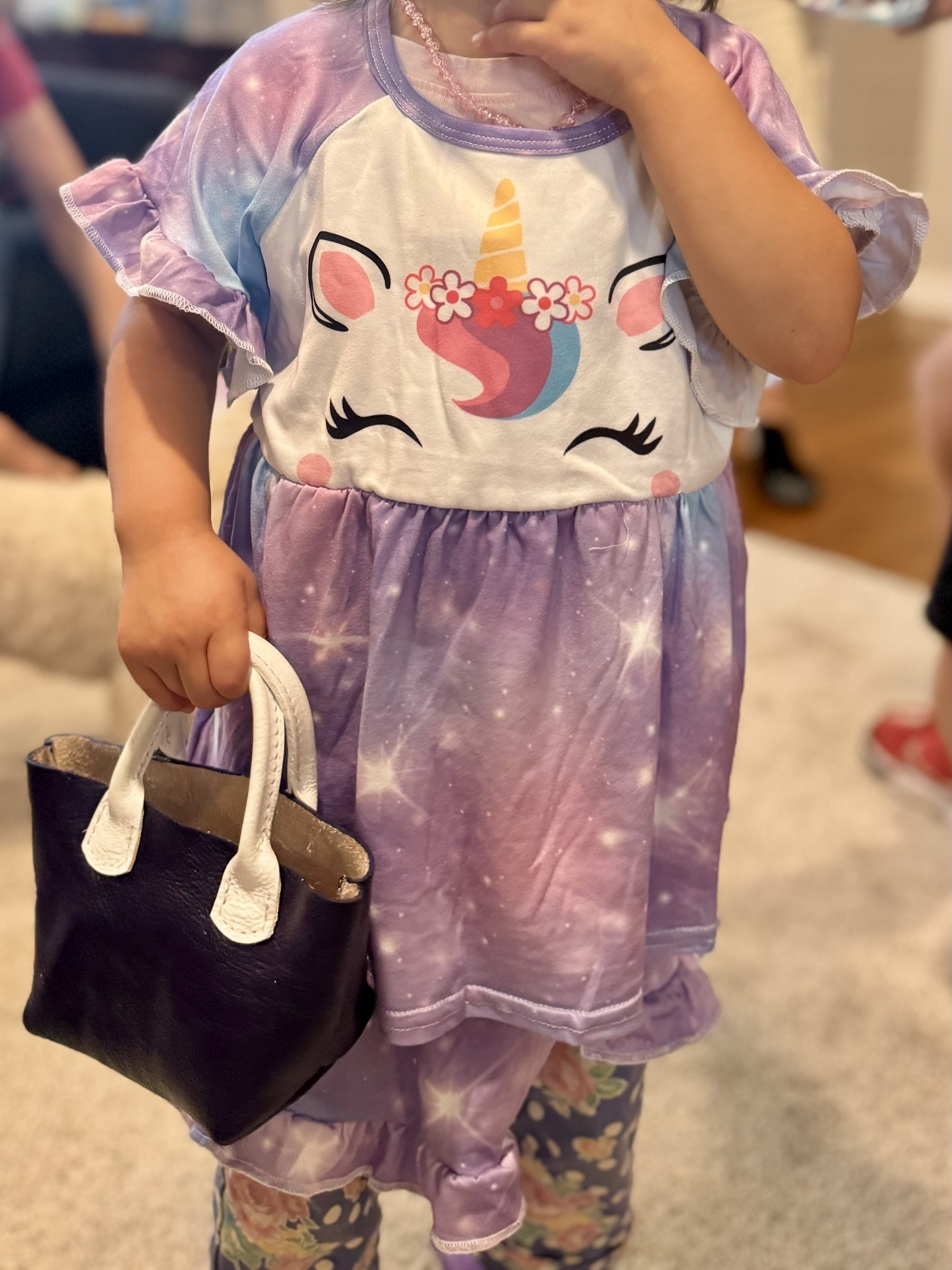 A child wearing a purple unicorn-themed dress and holding a small purple purse stands indoors.